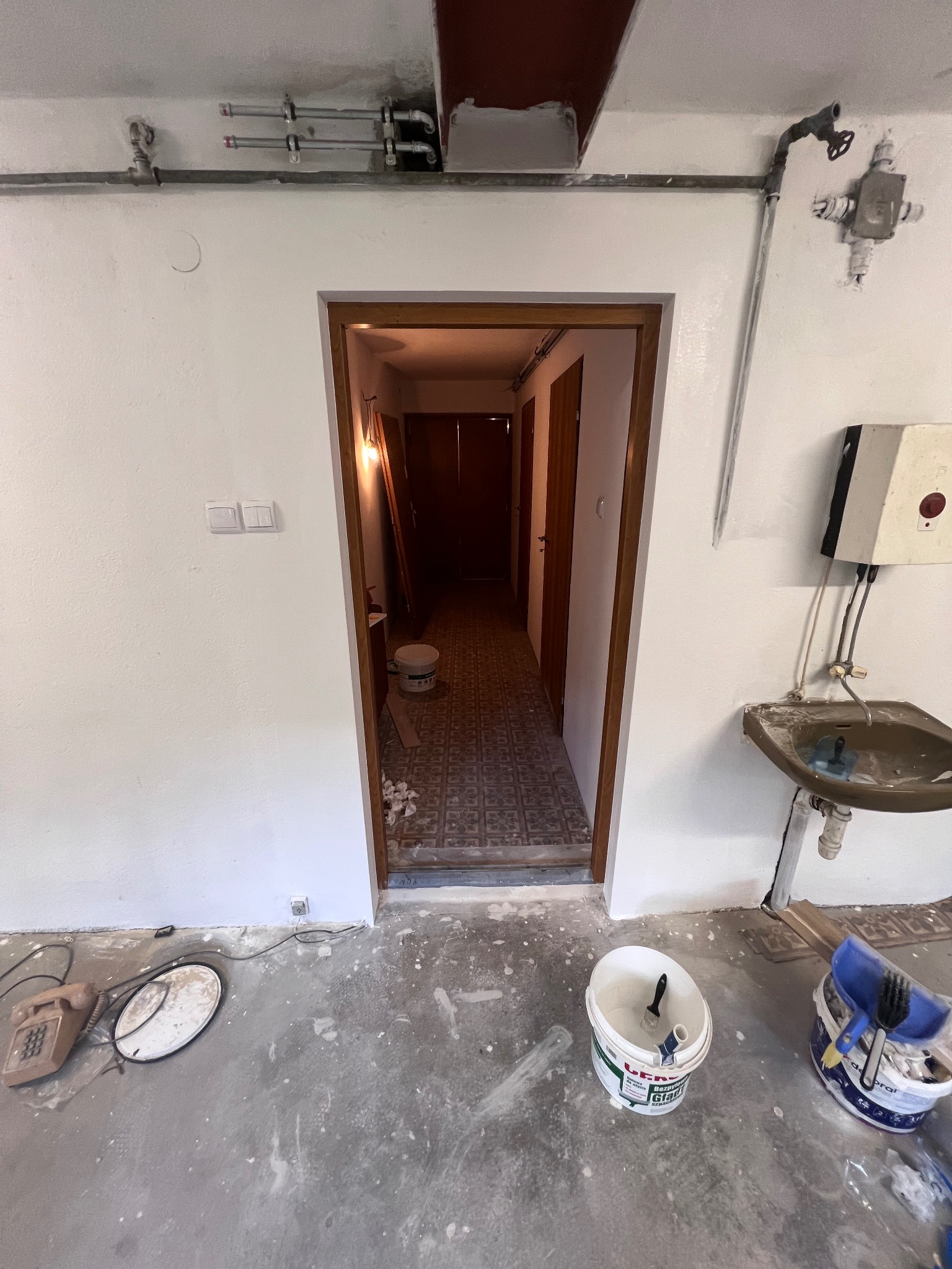 Wnętrze w trakcie malowania: ściany pokryte świeżą, białą farbą, widoczne narzędzia malarskie, puszki z farbą i niedokładności na podłodze. Remont w toku.