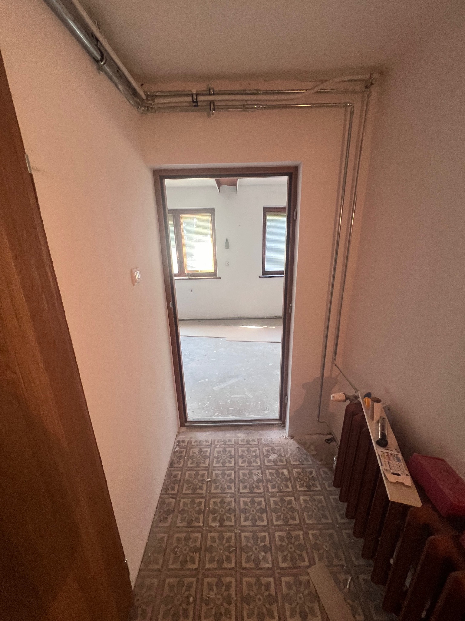 Wnętrze w trakcie remontu: widok na pokój przez otwarte drzwi, stara podłoga z wzorem, grzejnik z narzędziami, rury pod sufitem. Ściany przygotowane do malowania.