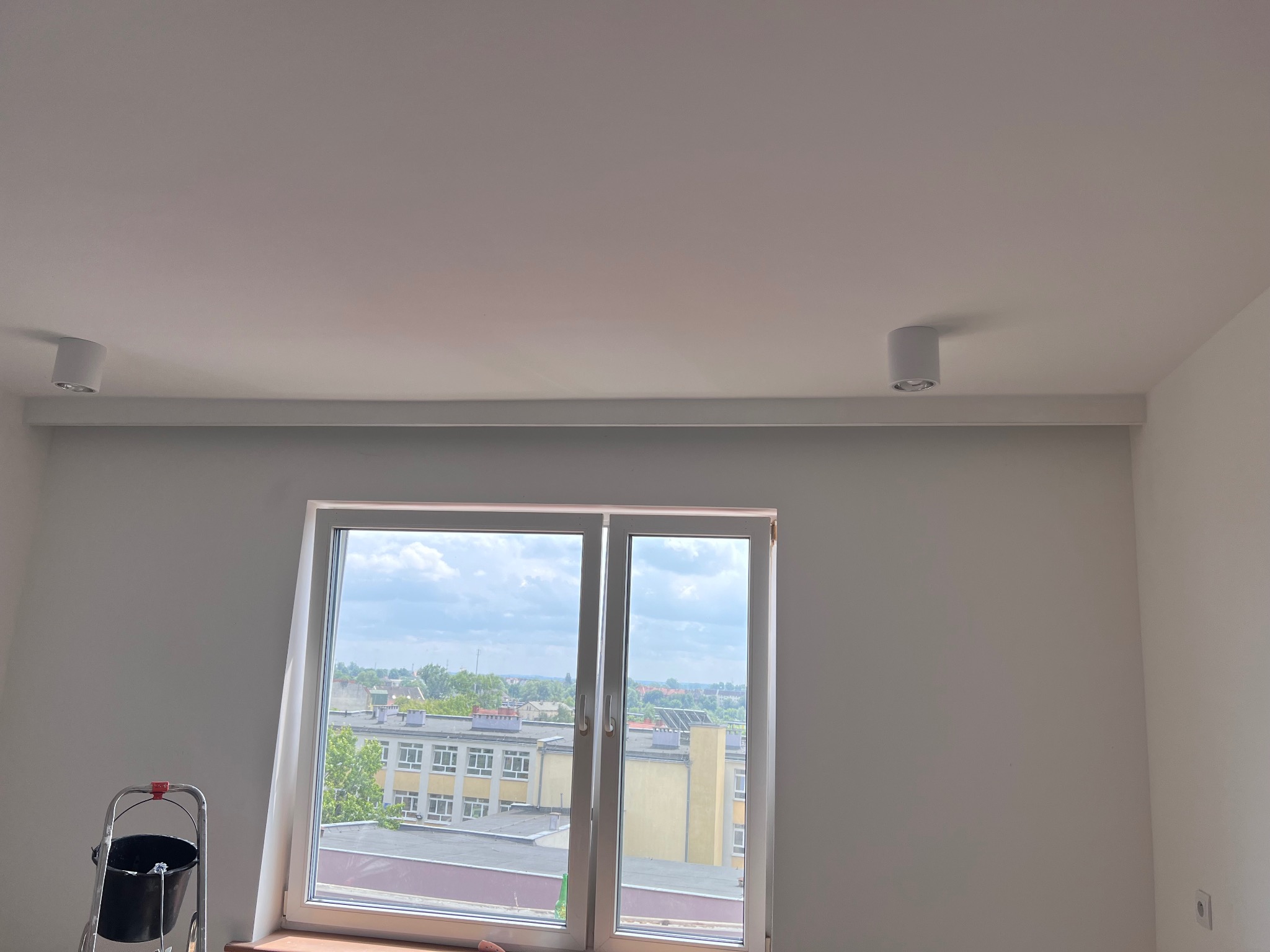 Białe wnętrze po malowaniu ścian z oknem i widokiem na miasto. Drabina malarska z wiadrem w rogu. Nowoczesne oświetlenie sufitowe punktowe.