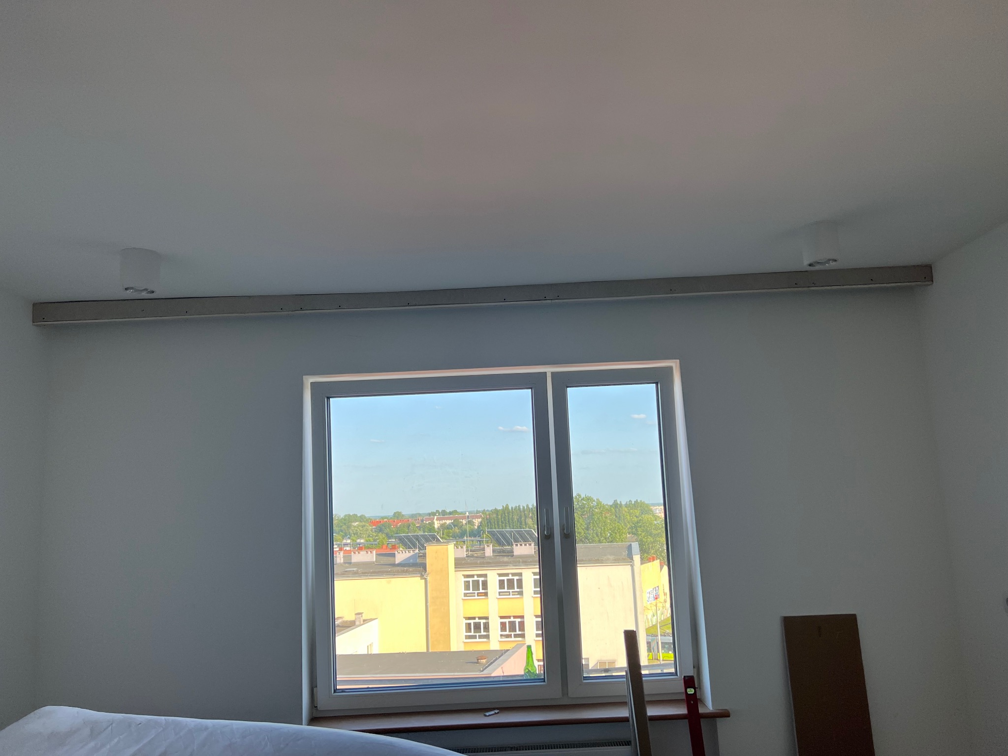 Wnętrze pokoju z oknem i zamontowaną listwą oświetleniową pod sufitem. Widoczne przygotowania do montażu oświetlenia. Świeżo pomalowane ściany.