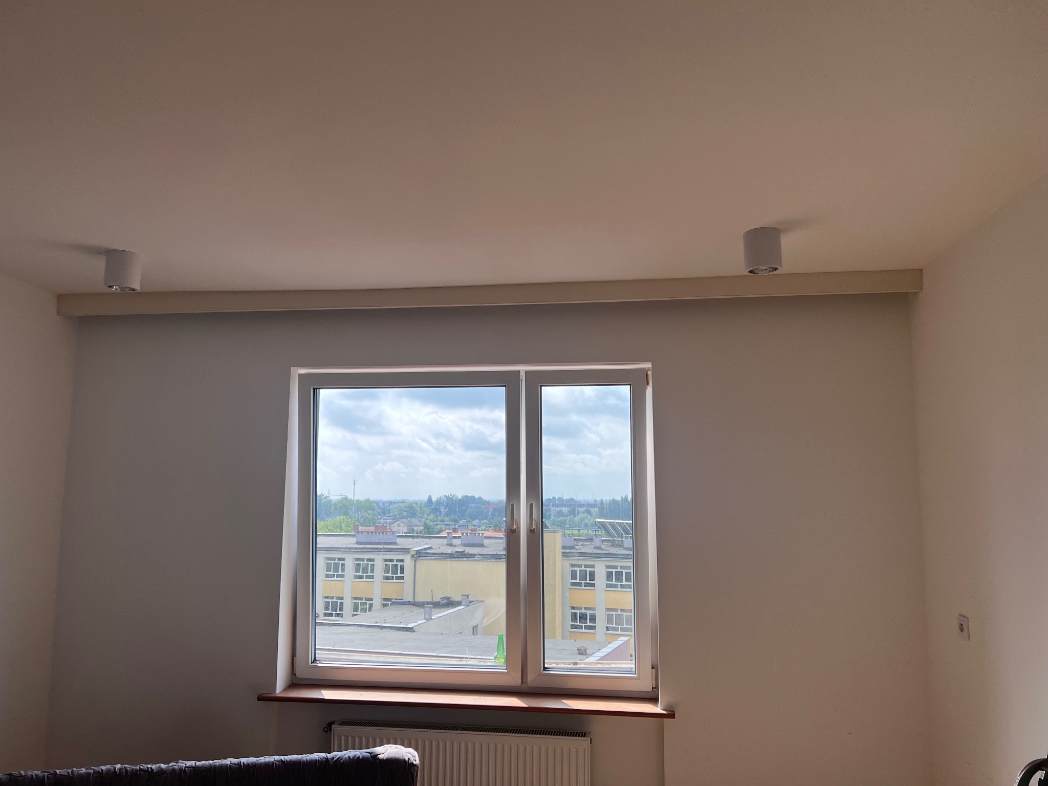 Jasne wnętrze z oknem na budynek szkolny. Dwa okrągłe, białe reflektory sufitowe. Świeżo pomalowane ściany w odcieniach bieli i beżu. Grzejnik pod oknem.