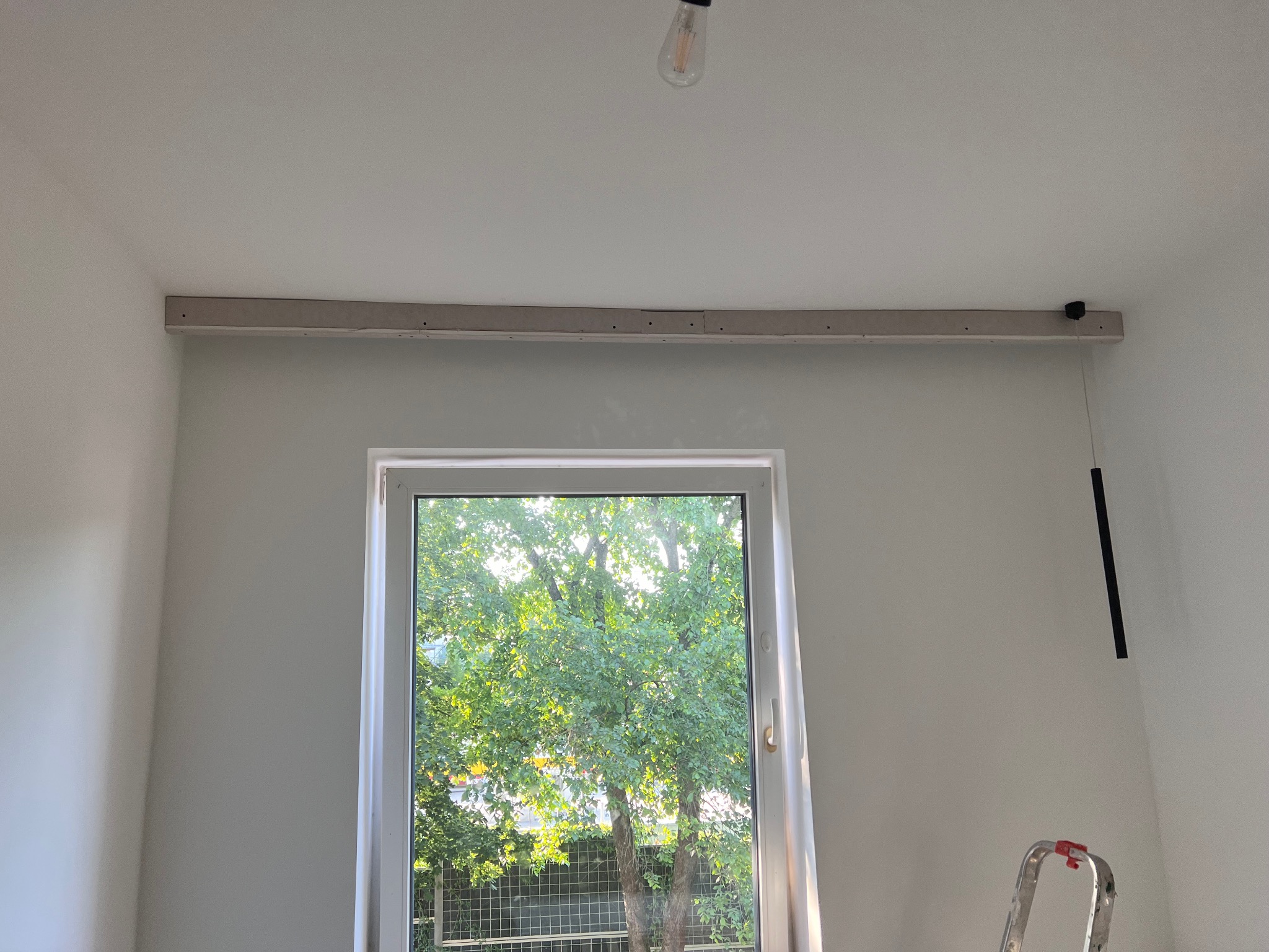 Wnętrze pokoju w trakcie wykończenia: zamontowana listwa sufitowa nad oknem, widoczna drabina i żarówka. Przez okno widać zieleń drzew.