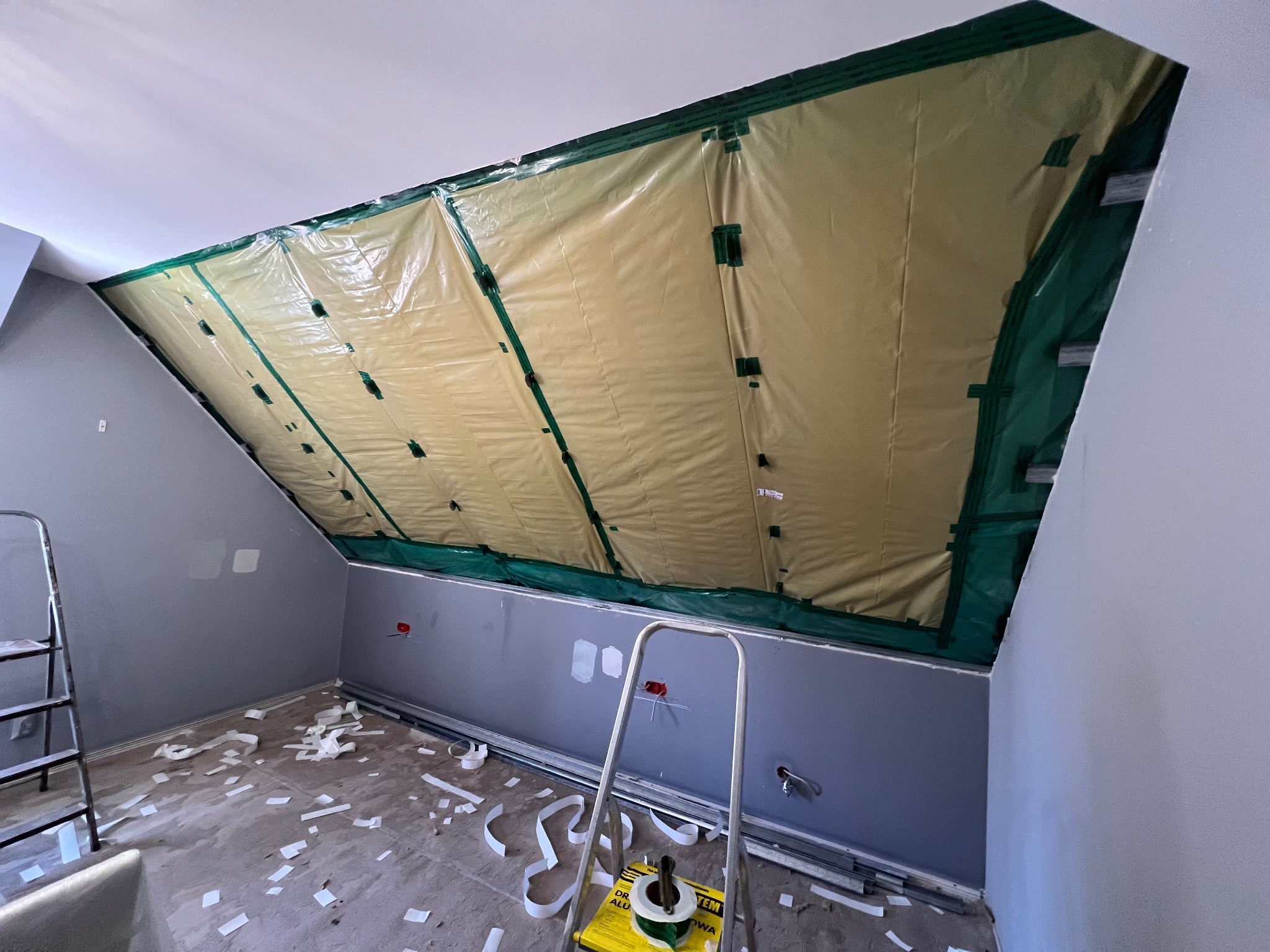 Poddasze w trakcie izolacji folią paroizolacyjną, widoczne drabiny i elementy montażowe na podłodze. Ściany pomalowane na szaro, sufit biały.