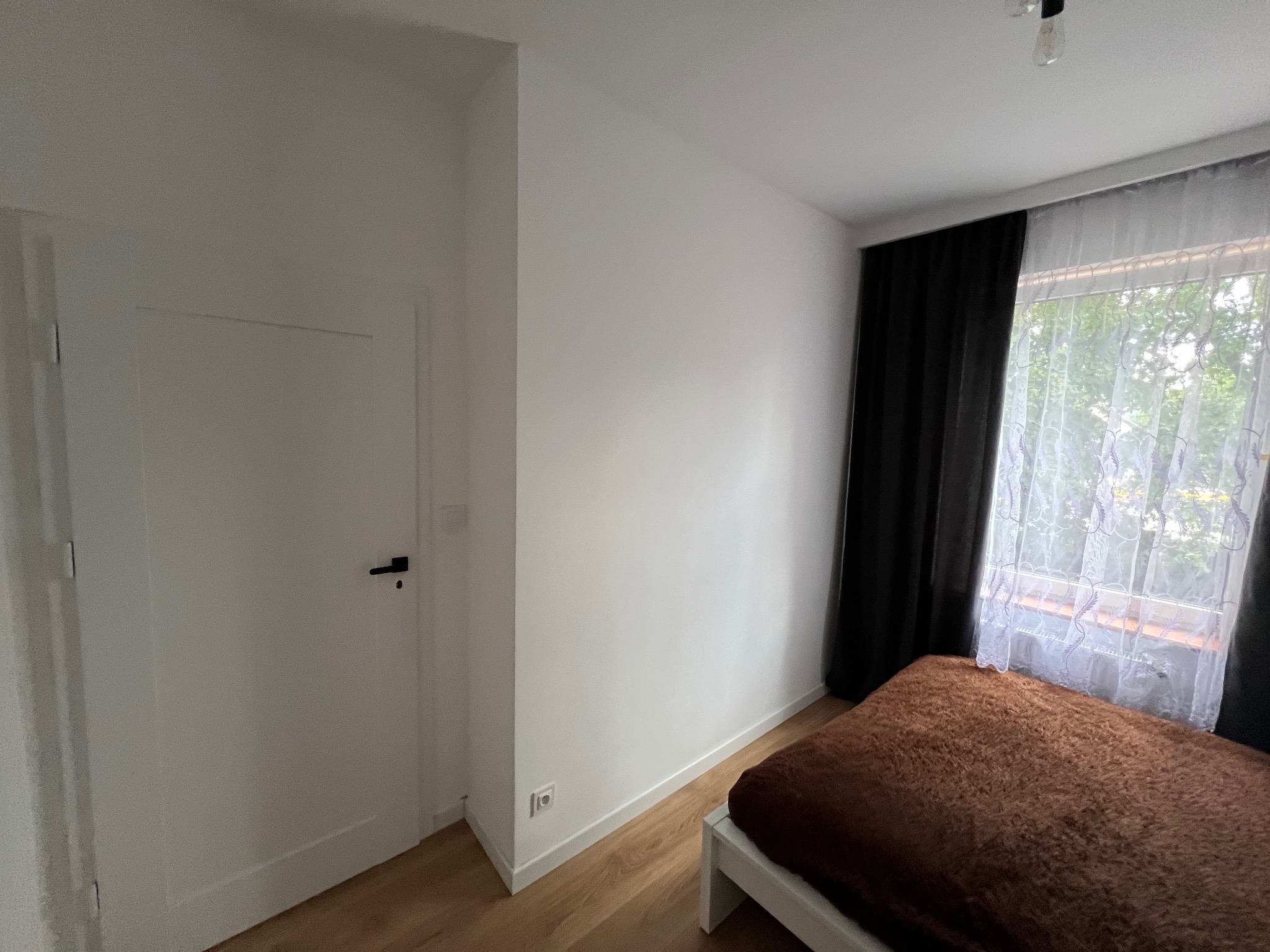 Pokój z białymi ścianami, drewnianą podłogą i łóżkiem z brązową narzutą przy oknie z czarnymi i białymi zasłonami. Widoczna biała, zamknięta  płycinowa drzwi z czarną klamką.
