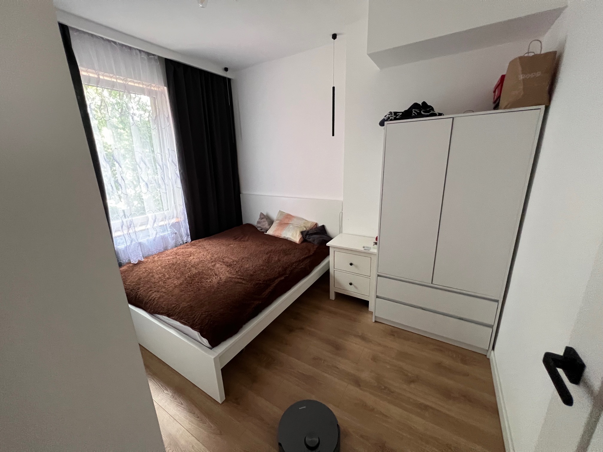 Ustawna sypialnia z drewnianą podłogą, białymi meblami i łóżkiem z brązową narzutą. Okno z firanką i zasłoną. Widoczny robot sprzątający.