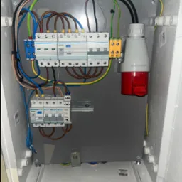 Szafa elektryczna z zainstalowanymi bezpiecznikami, przewodami i gniazdem siłowym. Widoczne elementy instalacji elektrycznej wewnątrz białej obudowy.