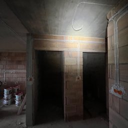 Instalacje Elektryczne - Wiktor Front - Surowy stan: widok wnętrza z instalacją elektryczną w budynku z cegły. Białe kable prowadzone po ścianach i suficie, puszki elektryczne w ścianach, brak wykończenia.
