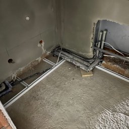 Instalacje Elektryczne - Wiktor Front - Wnętrze w trakcie remontu: surowe ściany z instalacjami elektrycznymi i hydraulicznymi, widoczne przewody i rury, betonowa wylewka podłogi w szarej tonacji.