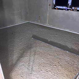 Instalacje Elektryczne - Wiktor Front - Wnętrze w trakcie wylewki podłogi: widoczna szlichta cementowa z listwami dylatacyjnymi przy ścianach. Okno z profilami zabezpieczającymi.