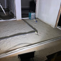 Instalacje Elektryczne - Wiktor Front - Układanie przewodów w podłodze przed wylewką. Widoczne białe rury i szary peszel. Na podłodze piasek. Sprzęt budowlany w tle. Etap prac instalacyjnych.