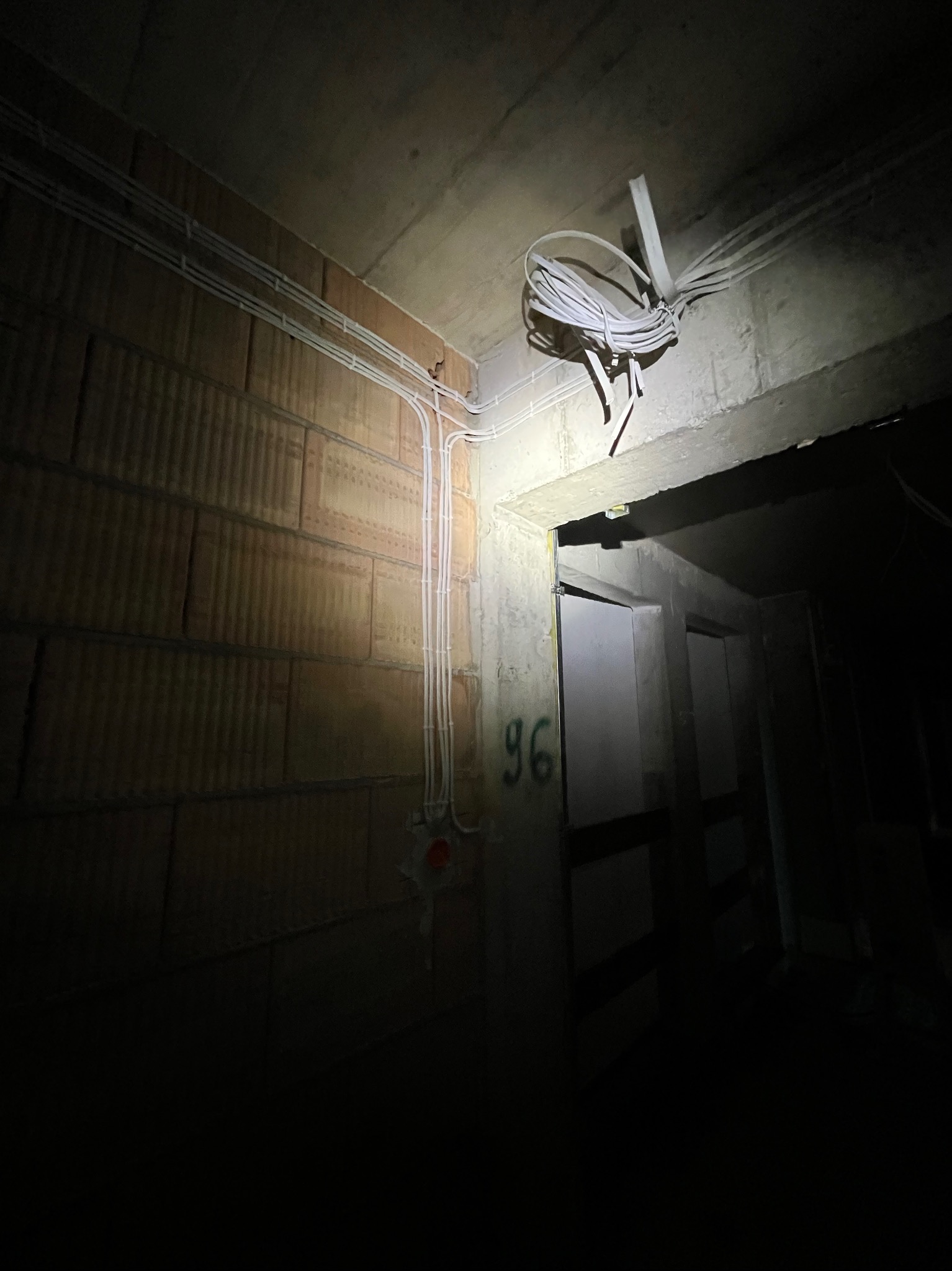 Białe kable elektryczne ułożone wzdłuż ceglanej ściany i betonowego sufitu w ciemnym pomieszczeniu. Część kabli zwisa w pętli. Widoczny fragment futryny drzwi.