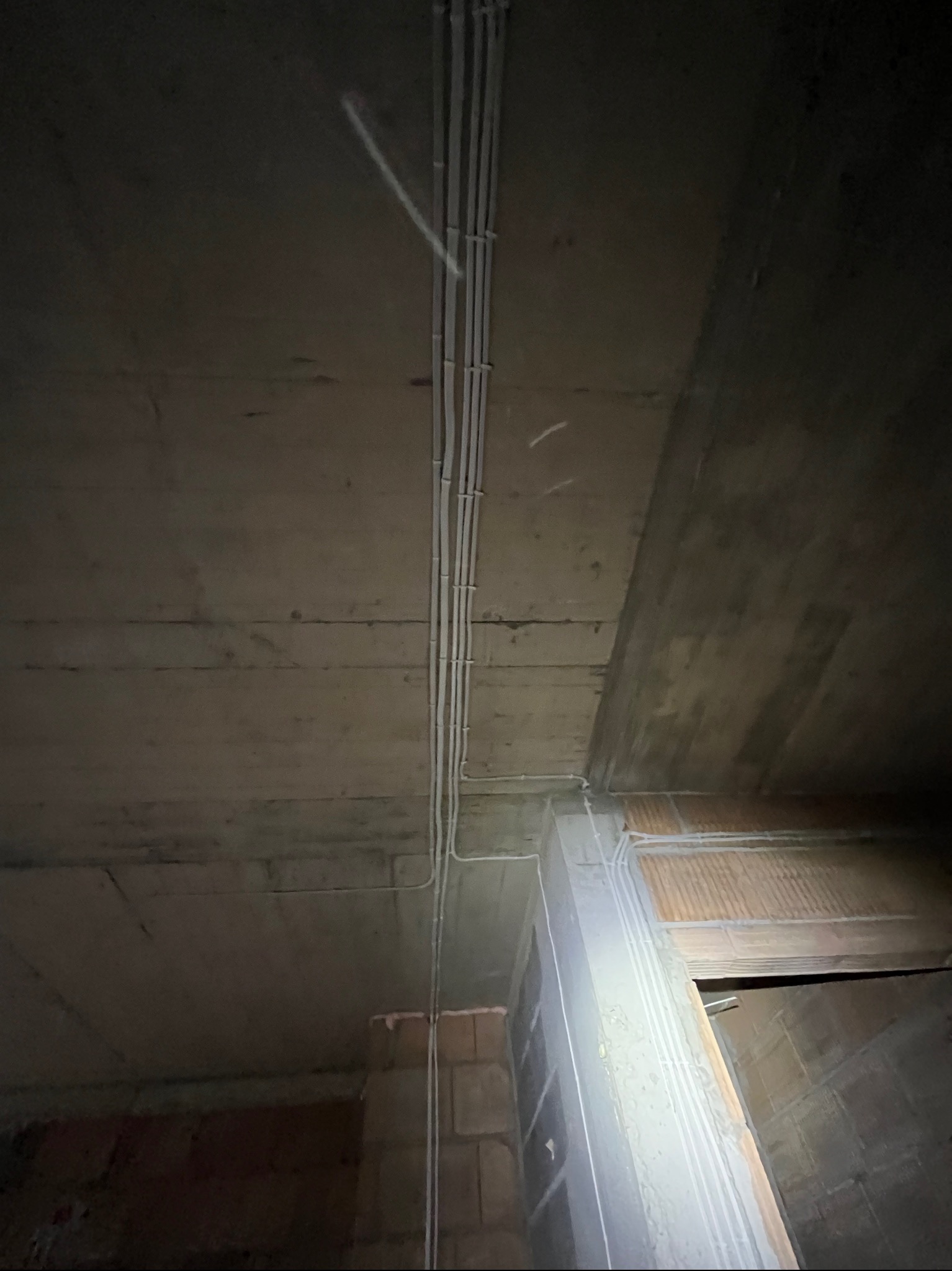 Białe kable elektryczne ułożone w rurkach na surowym betonowym suficie. Widoczne cegły i fragment otworu drzwiowego. Oświetlenie sufitowe.