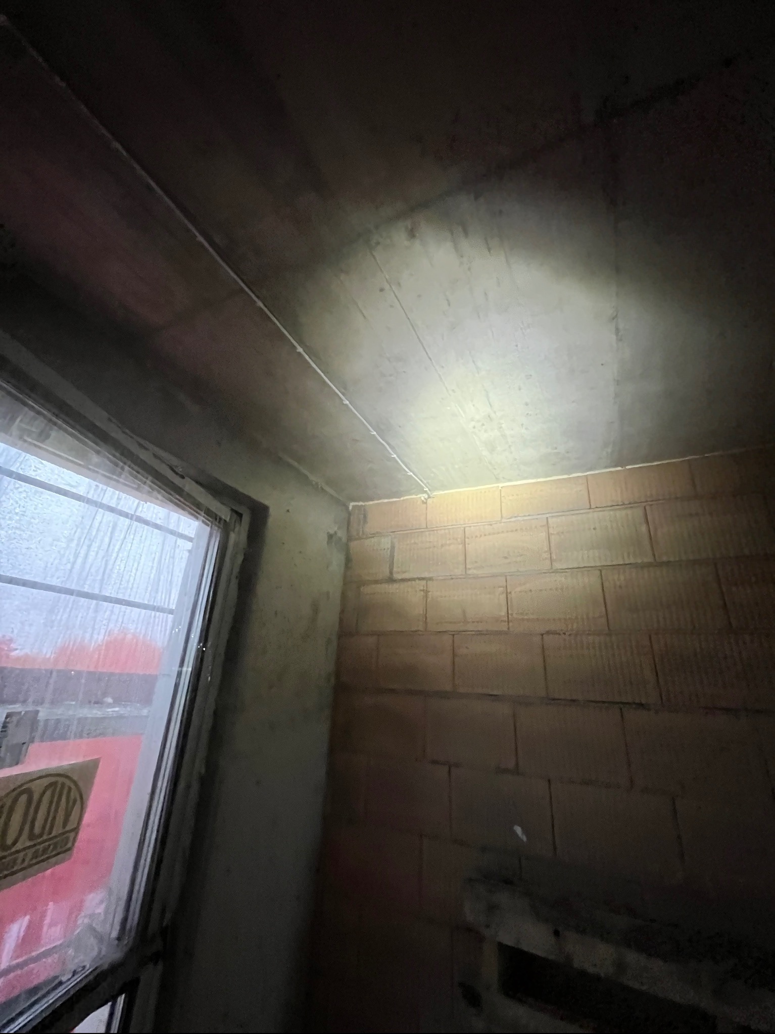 Wewnętrzne ujęcie surowego pomieszczenia z oknem, widoczne instalacje elektryczne pod sufitem i ściany z cegły. Surowy stan wykończenia.