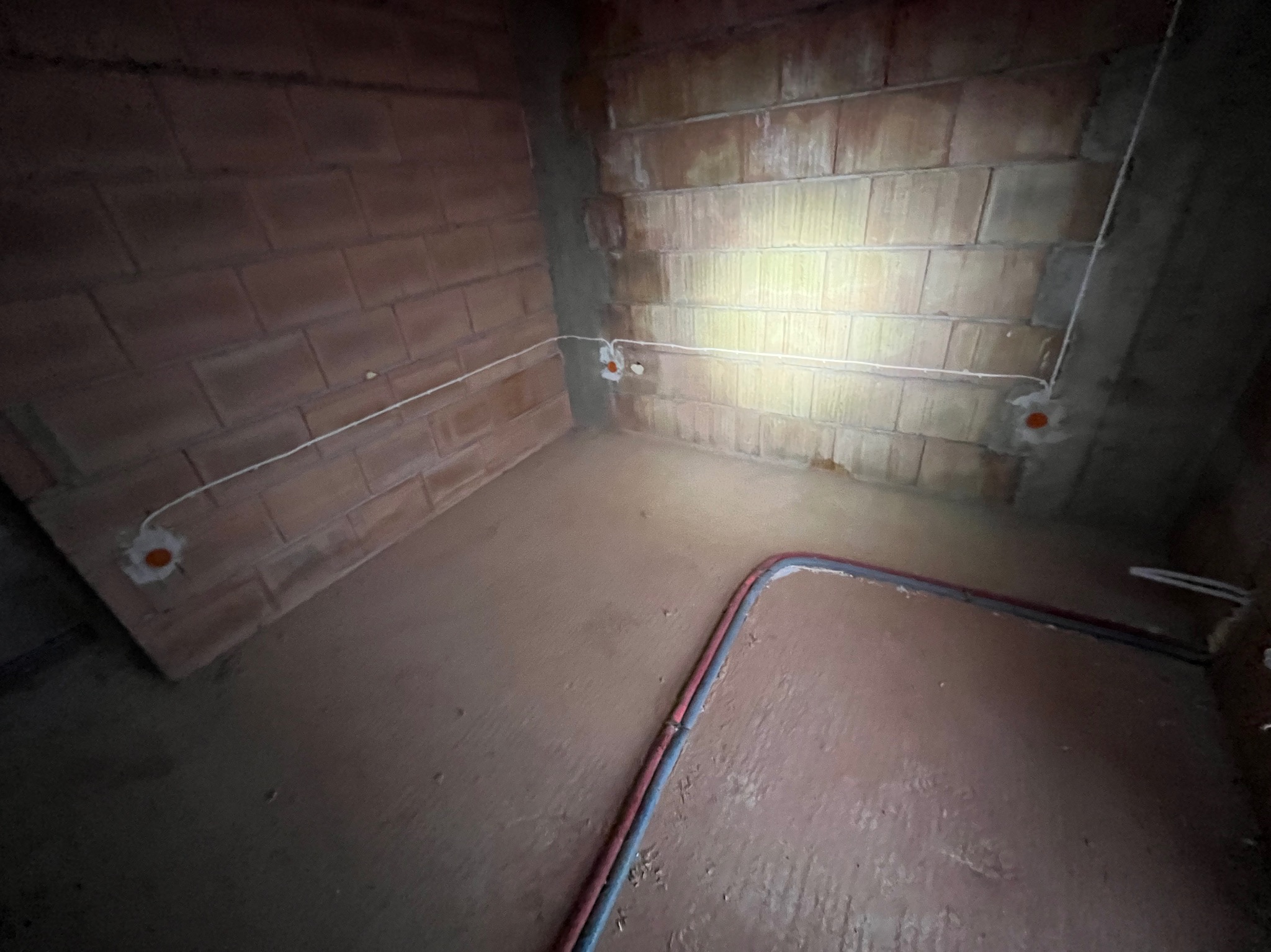 Surowy mur z cegły dziurawki z puszkami instalacyjnymi i przewodami. Podłoga wylewka. Instalacja elektryczna w trakcie realizacji w pomieszczeniu.