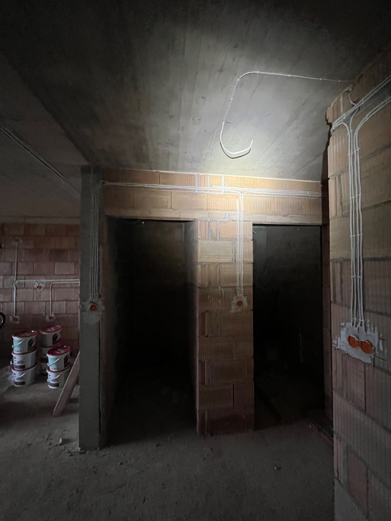 Surowy stan: widok wnętrza z instalacją elektryczną w budynku z cegły. Białe kable prowadzone po ścianach i suficie, puszki elektryczne w ścianach, brak wykończenia.