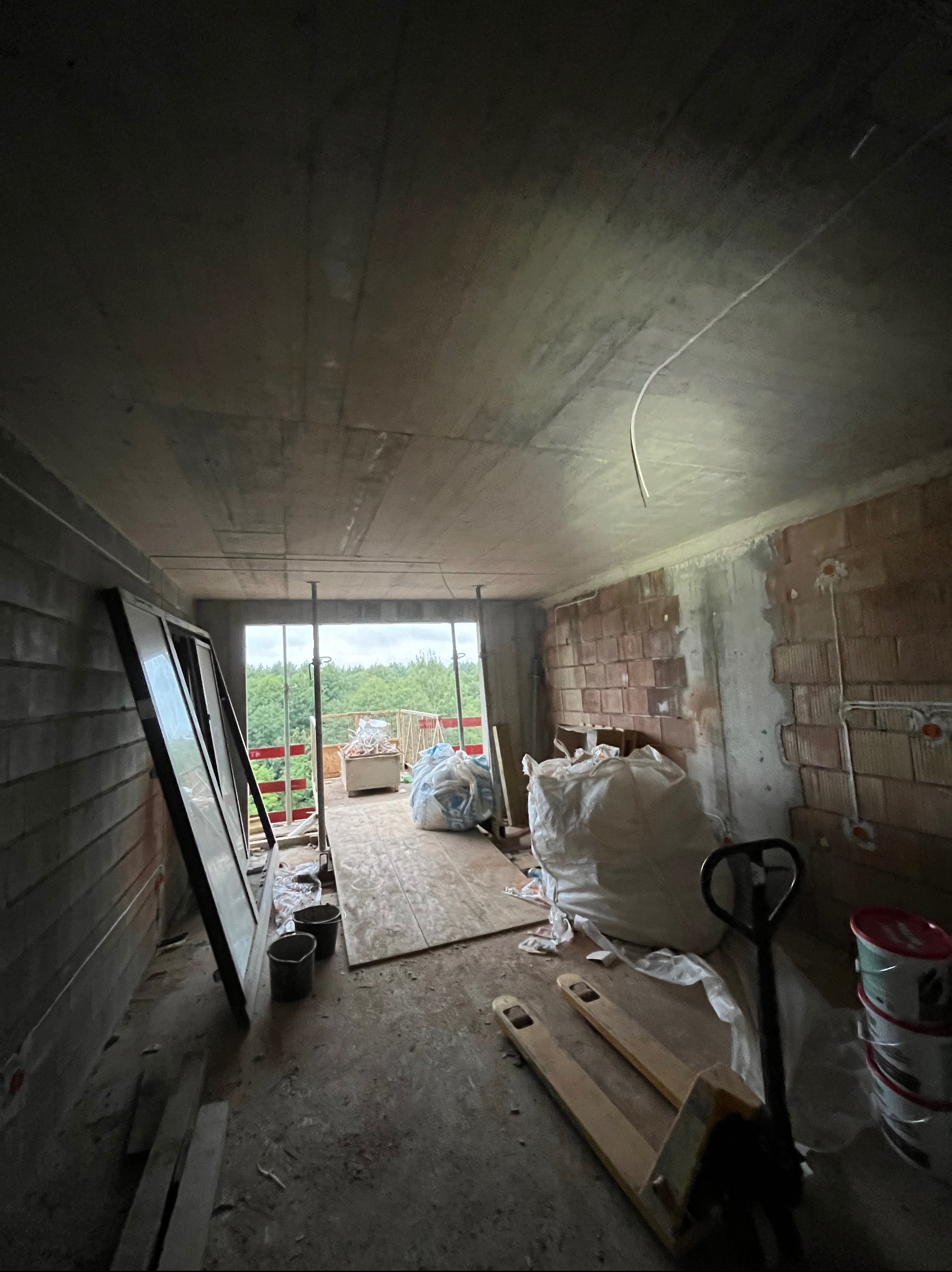 Wnętrze w trakcie budowy: surowe ściany z cegły, widoczne przewody elektryczne, worki z materiałami budowlanymi, paleciak. Przez okno widać zieleń.