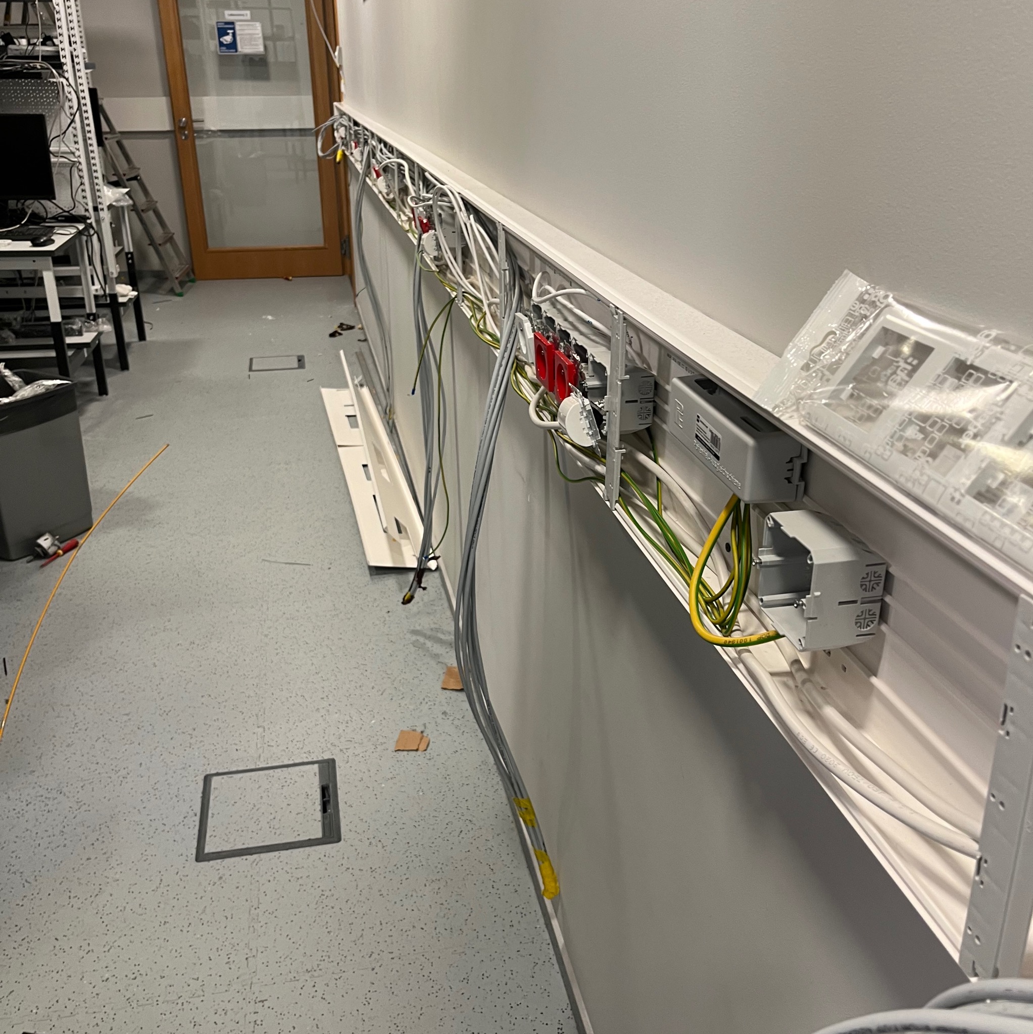 Montaż instalacji elektrycznej w korytarzu biurowym. Widoczne kable w korytkach, gniazda elektryczne i elementy montażowe. Prace w toku, widoczne narzędzia i materiały.