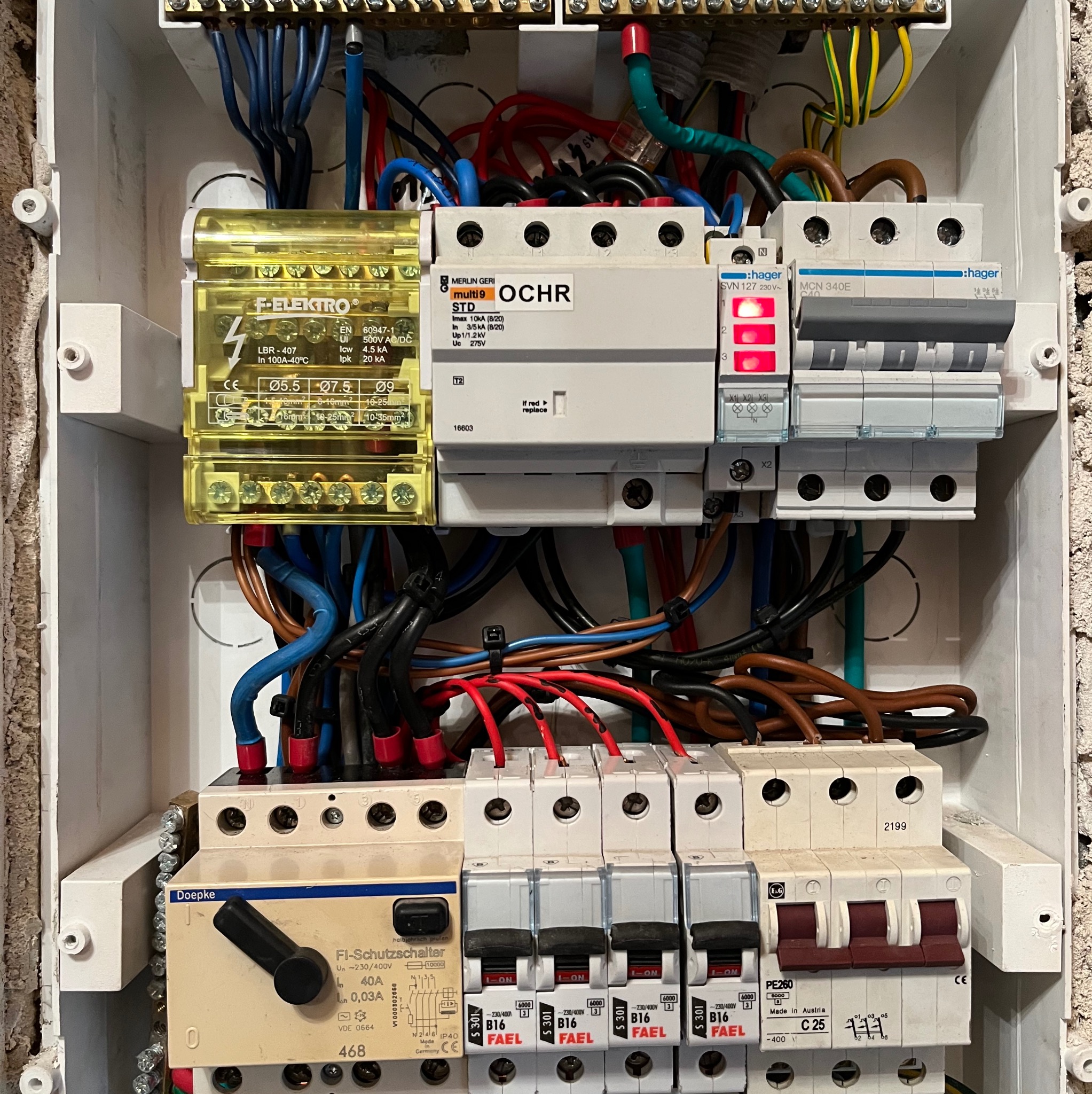 Rozdzielnica elektryczna z bezpiecznikami, przewodami i elementami sterującymi. Widoczne różne marki i modele urządzeń, w tym F-Elektro, Hager i Doepke.