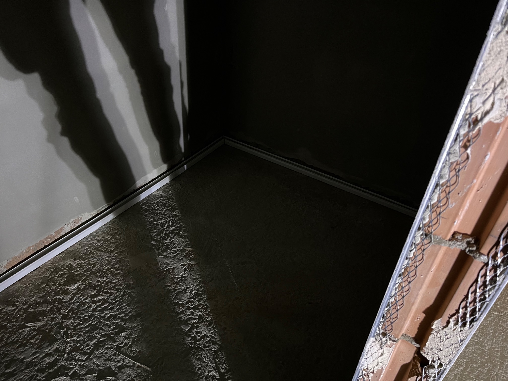 Surowa ściana w rogu pomieszczenia z listwą przypodłogową i fragmentem narożnika z siatką, przygotowana do dalszych prac wykończeniowych. Nierówna faktura podłoża.