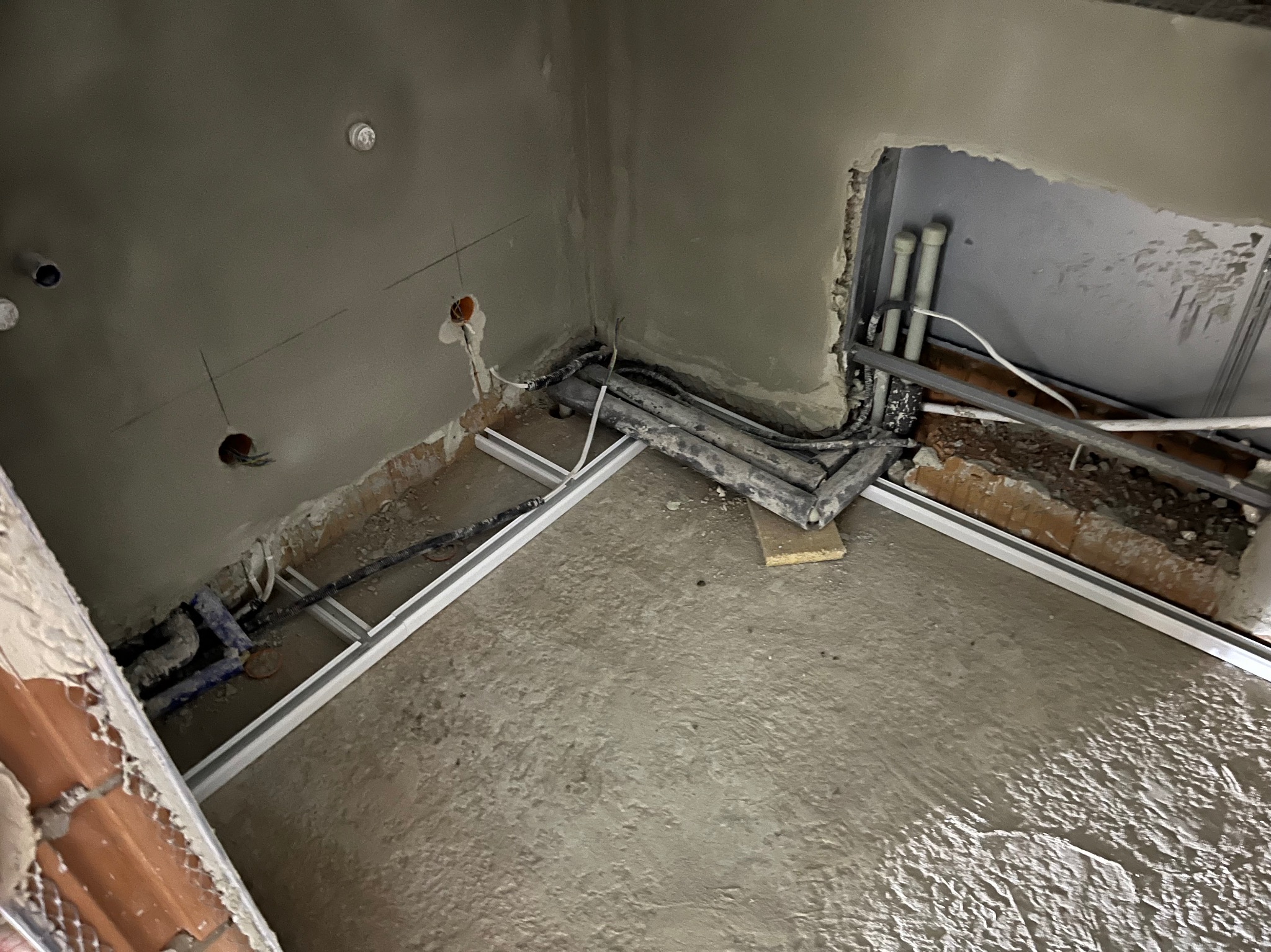 Wnętrze w trakcie remontu: surowe ściany z instalacjami elektrycznymi i hydraulicznymi, widoczne przewody i rury, betonowa wylewka podłogi w szarej tonacji.