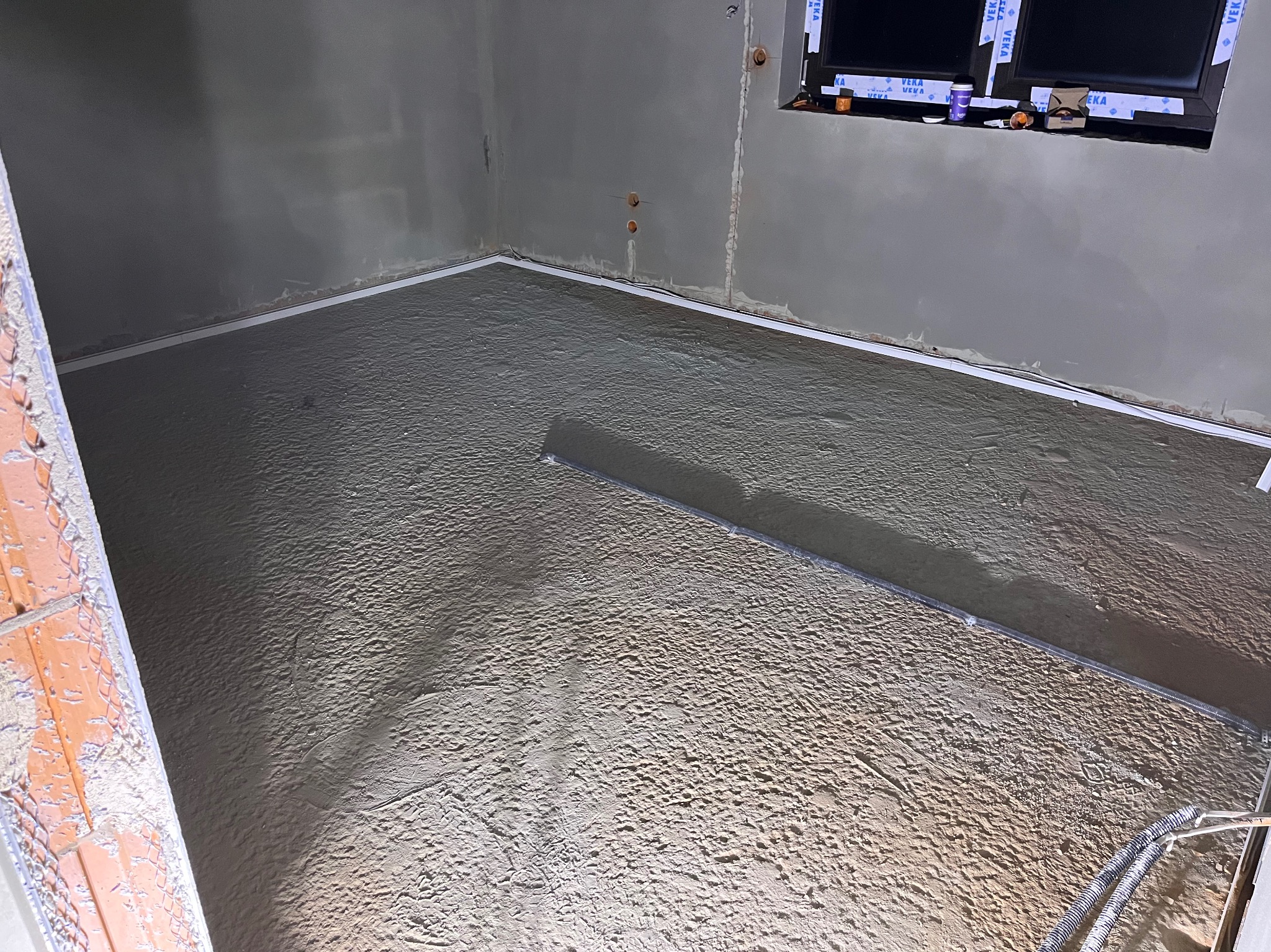 Wnętrze w trakcie wylewki podłogi: widoczna szlichta cementowa z listwami dylatacyjnymi przy ścianach. Okno z profilami zabezpieczającymi.