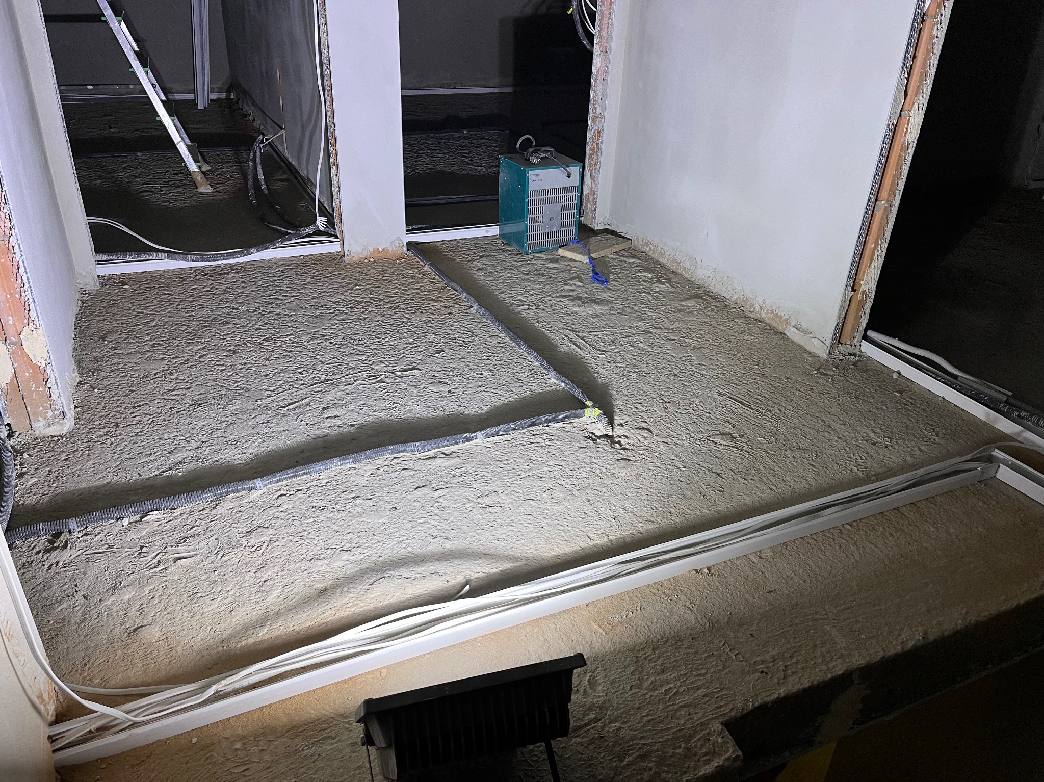 Układanie przewodów w podłodze przed wylewką. Widoczne białe rury i szary peszel. Na podłodze piasek. Sprzęt budowlany w tle. Etap prac instalacyjnych.