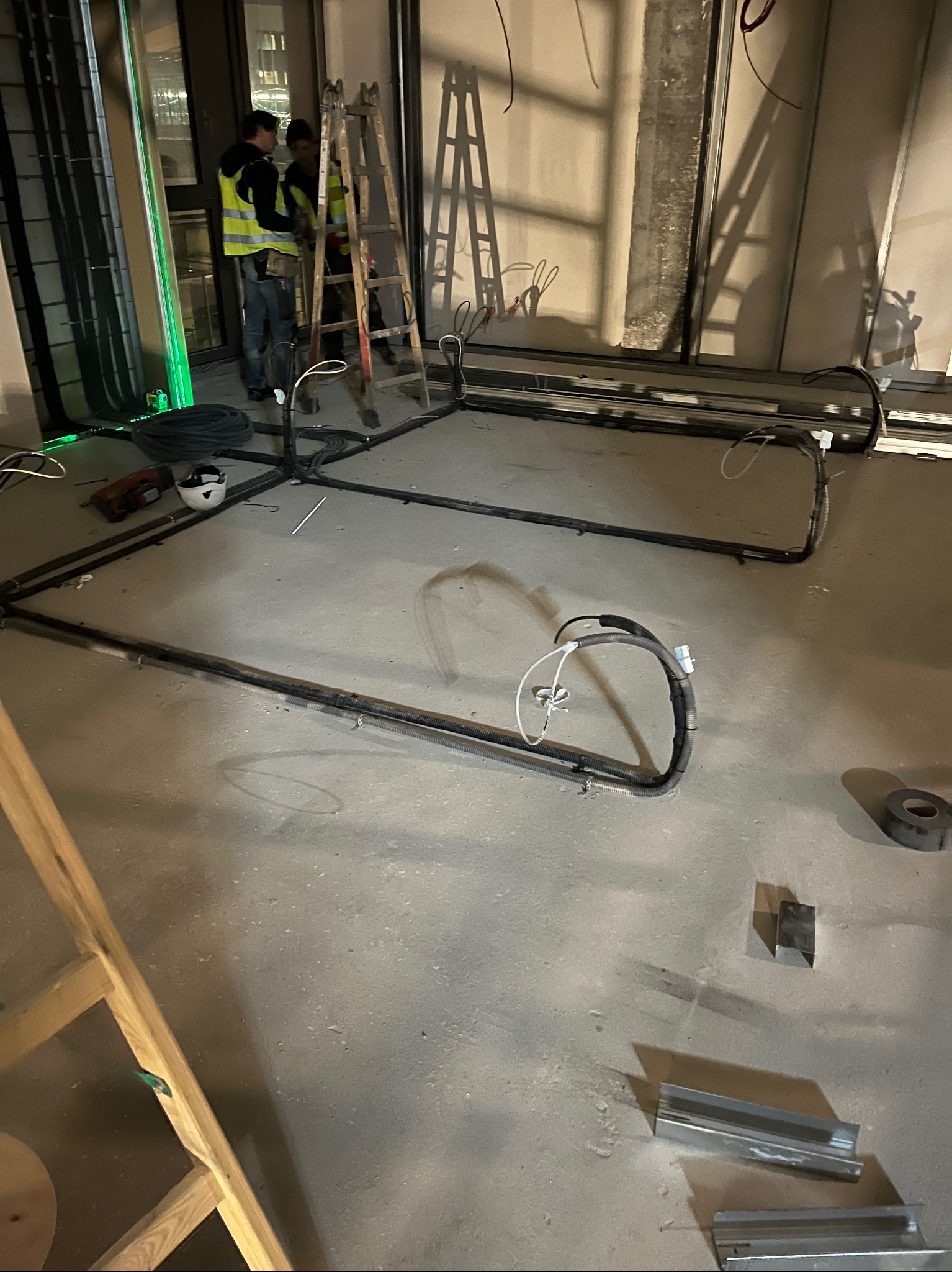 Montaż instalacji elektrycznej w nowym budynku. Widoczne kable ułożone na podłodze, drabina i pracownicy w odblaskowych kamizelkach. Elementy instalacyjne na posadzce.