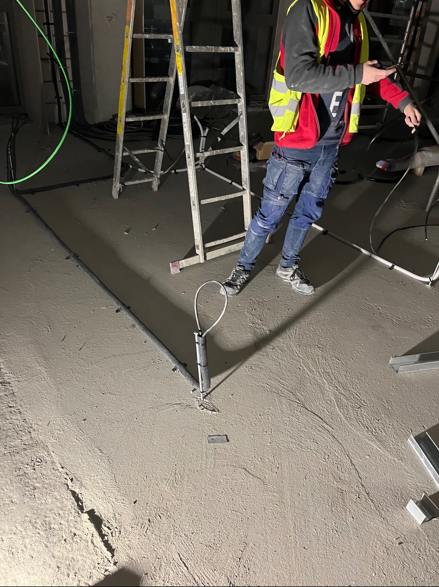 Elektryk w kamizelce BHP, instaluje przewody w podłodze. Widoczna drabina, narzędzia i kable. Szara podłoga, surowy stan. Praca w toku, widok z góry.