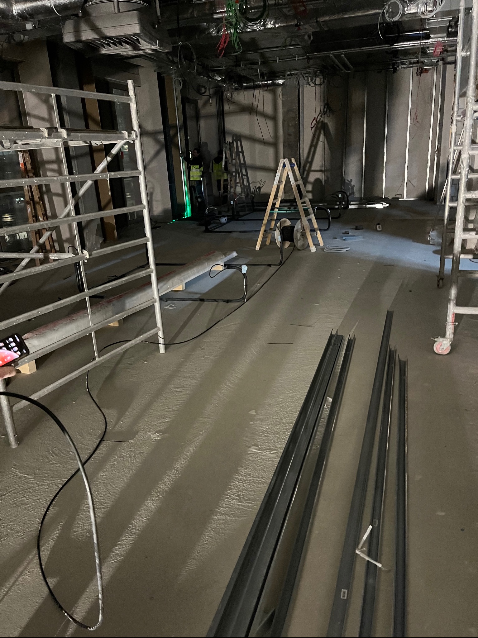 Wnętrze w trakcie prac instalacyjnych: widoczne kable, drabiny, profile metalowe i pracownicy w oddali. Surowy beton i instalacje pod sufitem.