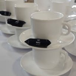 Anna Brembor B&G - Białe filiżanki z czarnymi czekoladkami na białych spodkach, ustawione w rzędach, przygotowane do serwowania, z bliska, w jasnym oświetleniu.