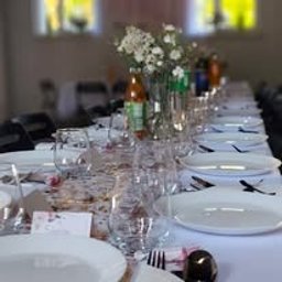 Anna Brembor B&G - Uroczysty stół z białymi talerzami, szkłem i kwiatami. Dekoracje w odcieniach bieli i złota. Widoczne napoje w szklanych butelkach. Przygotowania do przyjęcia.