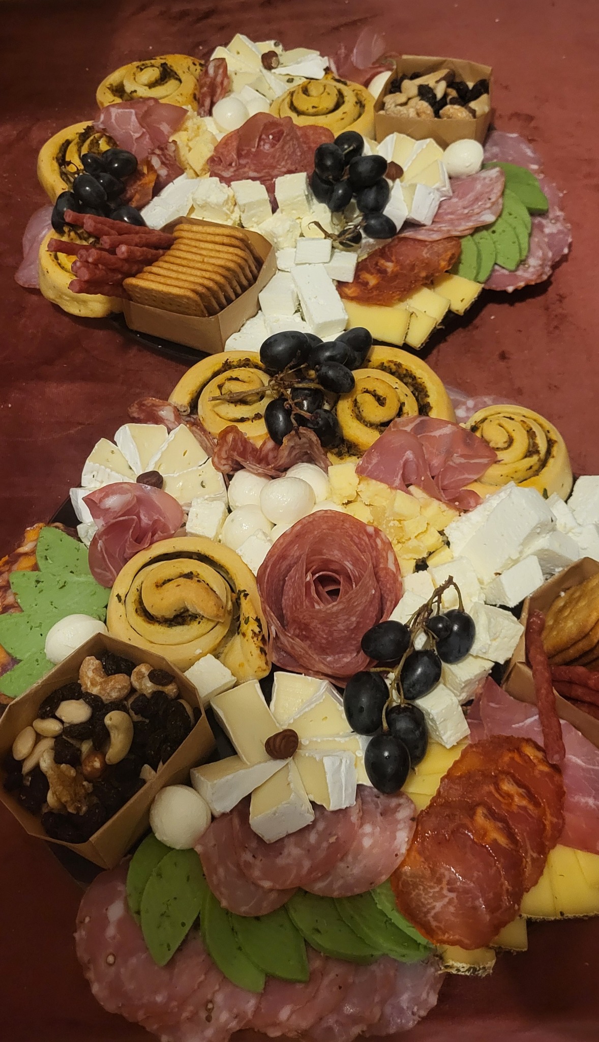 Dwa okrągłe półmiski z wykwintnymi przekąskami: sery, wędliny, rolady z pesto, owoce i orzechy, tworzące apetyczną kompozycję na ciemnoczerwonym tle.