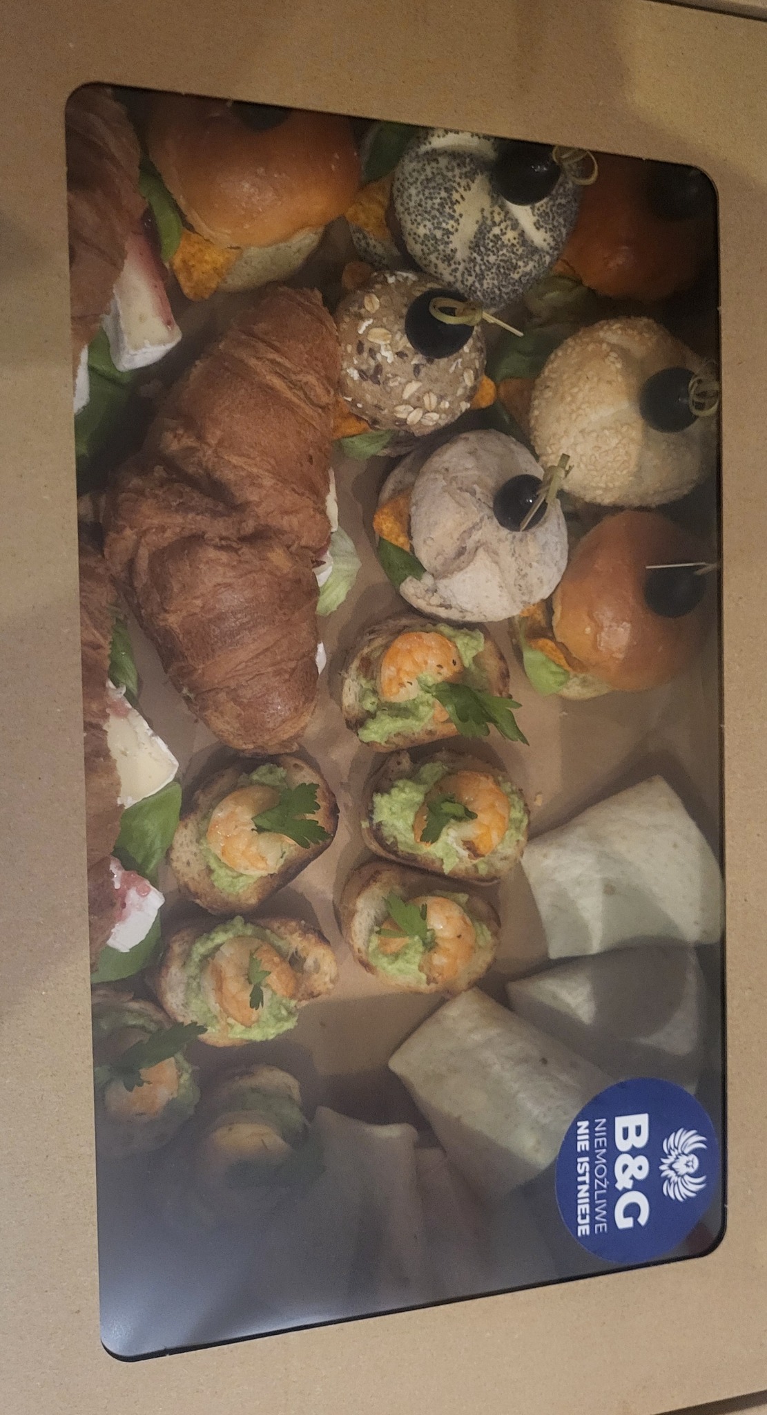 Różnorodne przekąski w kartonowym pudełku: mini kanapki z krewetkami, croissanty, burgery z oliwkami. Widoczny narożnik pudełka z naklejką firmy.