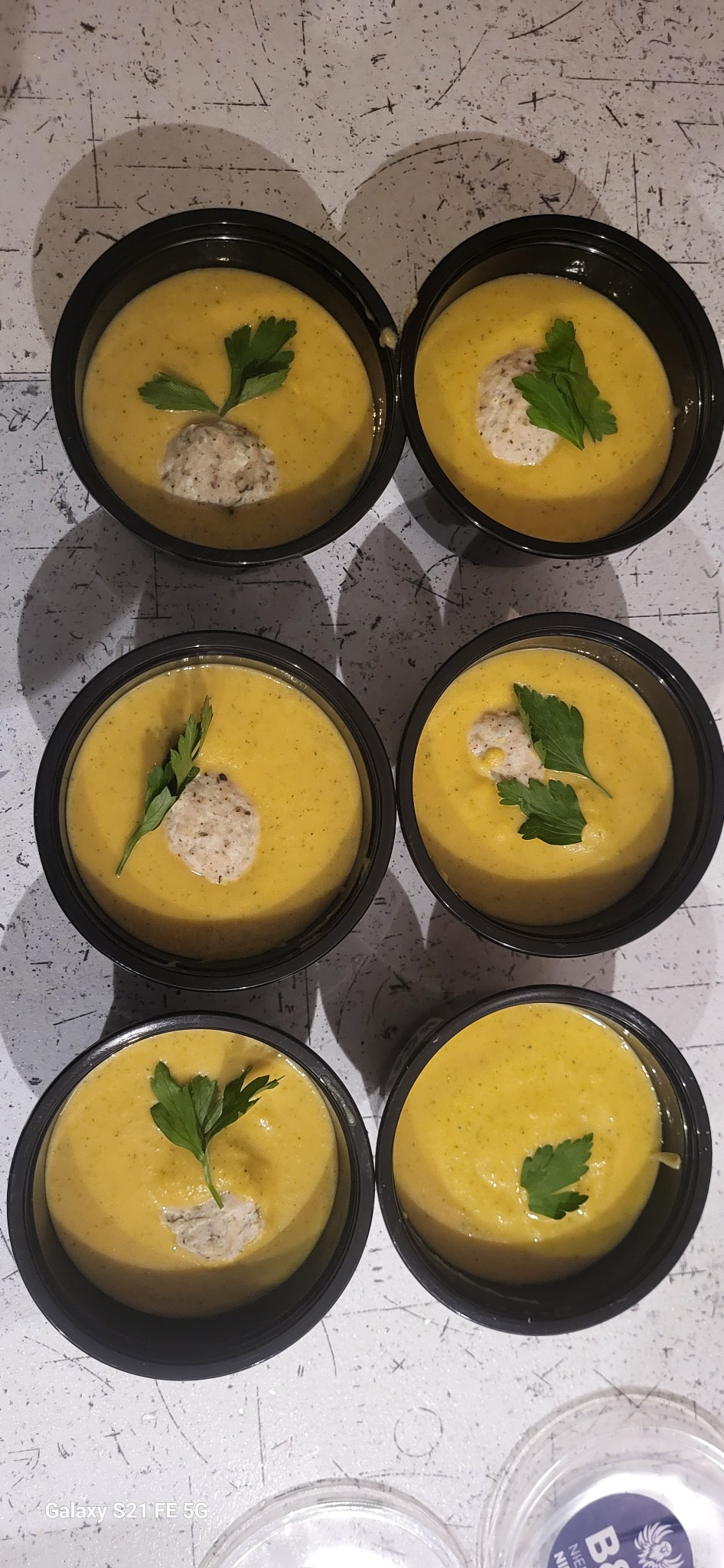 Sześć porcji kremowej zupy dyniowej w czarnych miseczkach, udekorowanych kleksem i zieloną pietruszką, na jasnym tle z widocznymi cieniami.