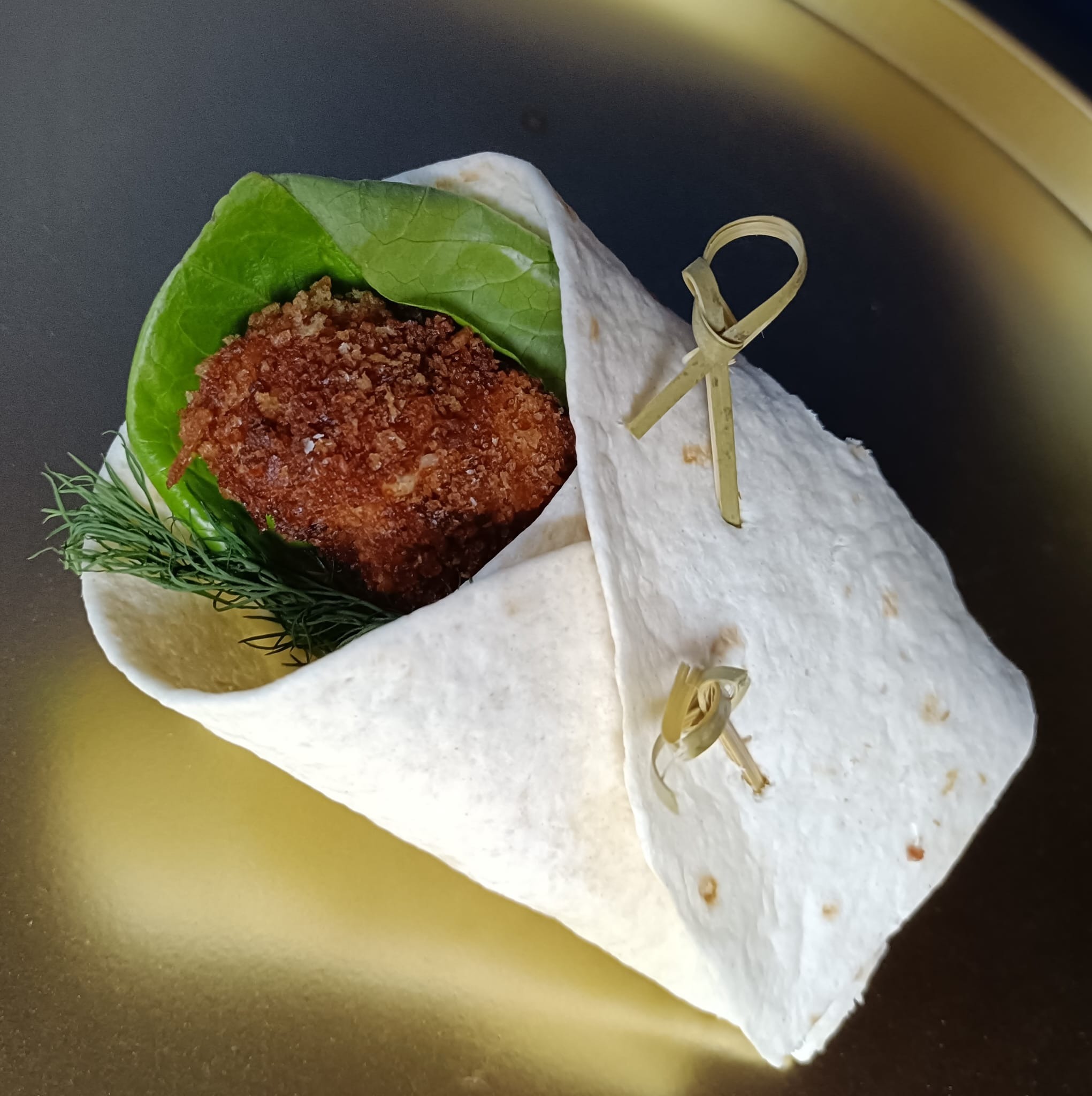 Tortilla wrap z kotletem w panierce, sałatą i koperkiem, spięta ozdobnymi wykałaczkami. Całość na złotym talerzu, w ciepłym oświetleniu.