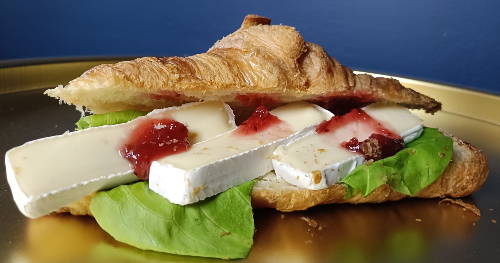 Kulinarna kompozycja: croissant z serem brie, dżemem i zielonymi liśćmi na złotym talerzu, na tle granatowej ściany. Delikatne światło podkreśla teksturę pieczywa.