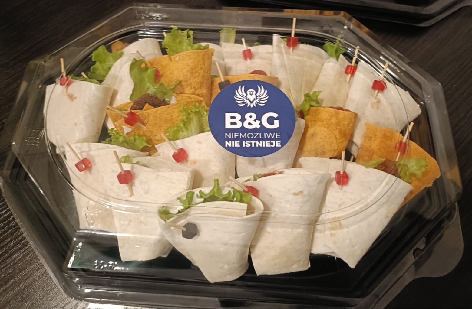 Tortilla wrap przekąski z sałatą i dekoracyjnymi wykałaczkami w przezroczystym opakowaniu cateringowym z logo firmy B&G, gotowe do serwowania na imprezie.