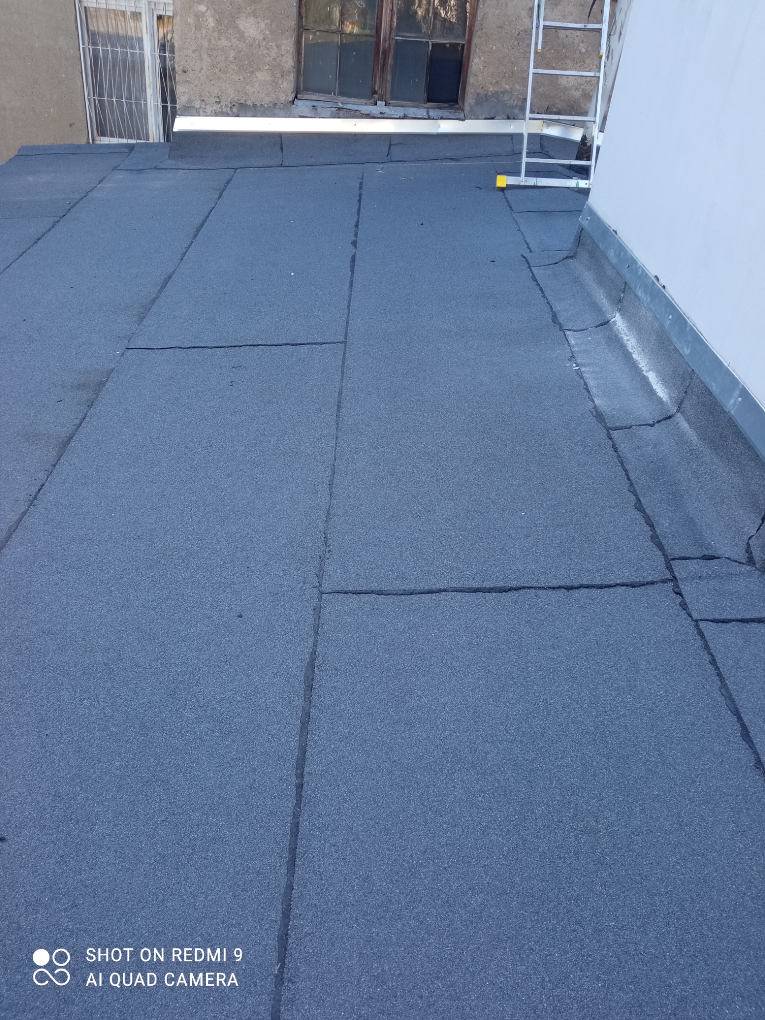 Szczelny, ciemnoszary dach pokryty papą termozgrzewalną z widocznymi łączeniami, drabina oparta o ścianę budynku w tle.