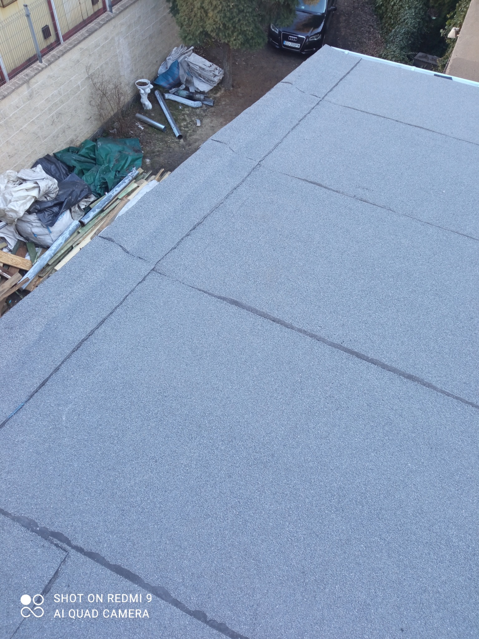 Szary, płaski dach pokryty papą termozgrzewalną, widok z góry pod kątem. W tle zaparkowany czarny samochód i elementy budowlane.