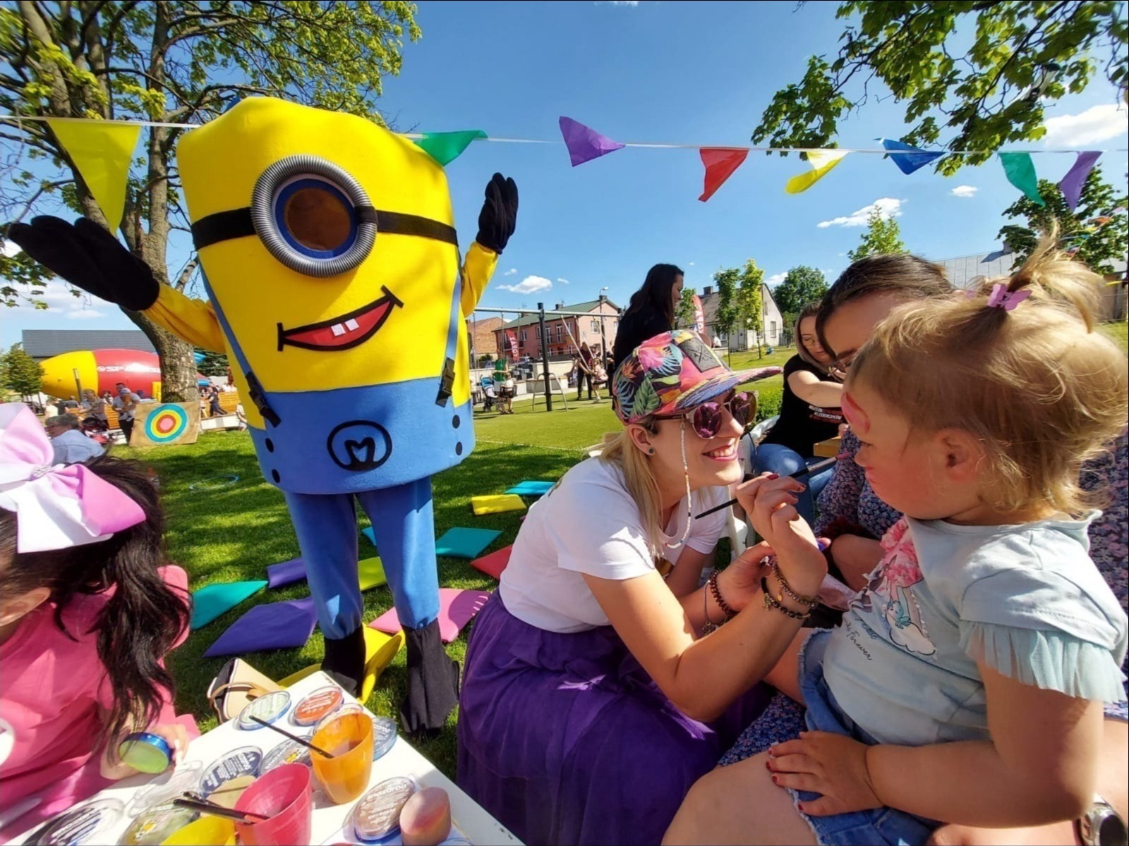 Animator maluje buzię dziecka na zielonej trawie, w tle Minionek i kolorowe dekoracje. Radosna atmosfera imprezy plenerowej dla dzieci.