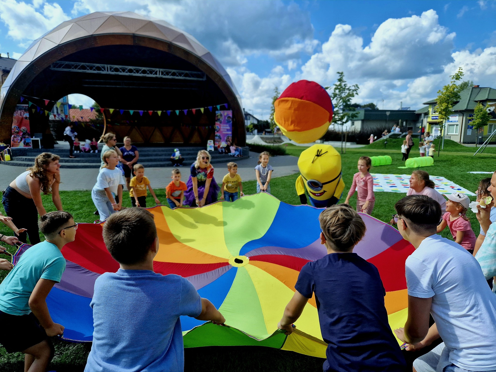 Dzieci trzymające kolorową chustę animacyjną na tle sceny plenerowej i animatorów w kostiumach. Radosna zabawa na imprezie dla dzieci.