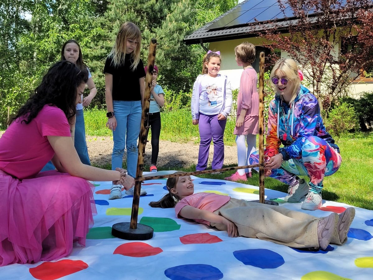 Zabawa Limbo na trawie: dzieci i animatorka w kolorowym stroju. Dziewczynka leży na macie do twistera, podnoszona poprzeczka. Radosna atmosfera.