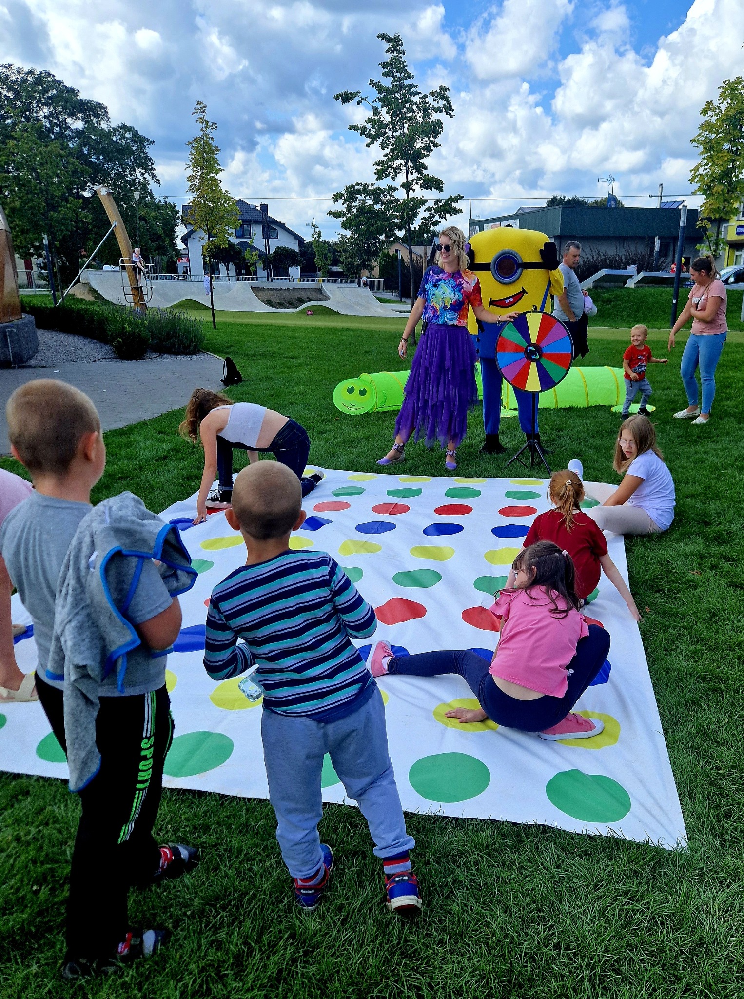 Animator w kostiumie Minionka prowadzi grę Twister na trawie z dziećmi. W tle dmuchany tunel, plac zabaw i osoby dorosłe.