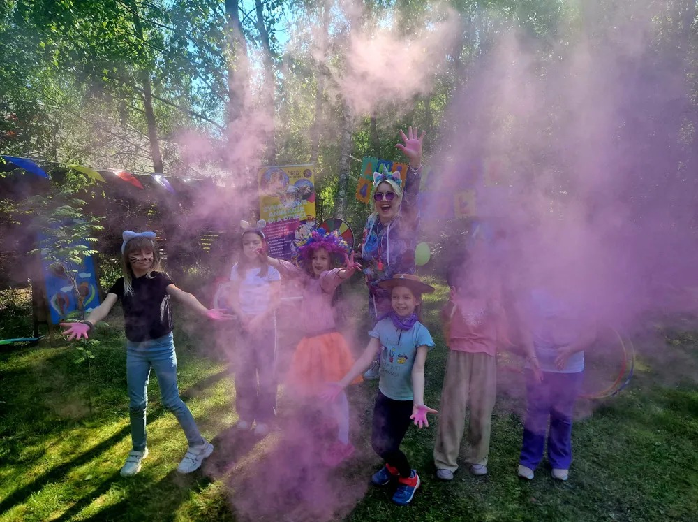 Grupa dzieci i animatorka w plenerze, rzucają kolorowym proszkiem podczas zabawy Holi. Kolorowe stroje, uśmiechy, tęczowa chmura proszku.