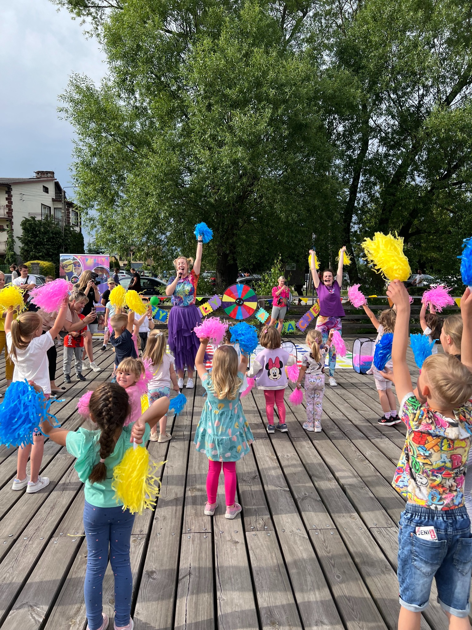 Animator z mikrofonem prowadzi grupę dzieci z kolorowymi pomponami na drewnianym podeście, w tle drzewa i budynki. Radosna atmosfera.
