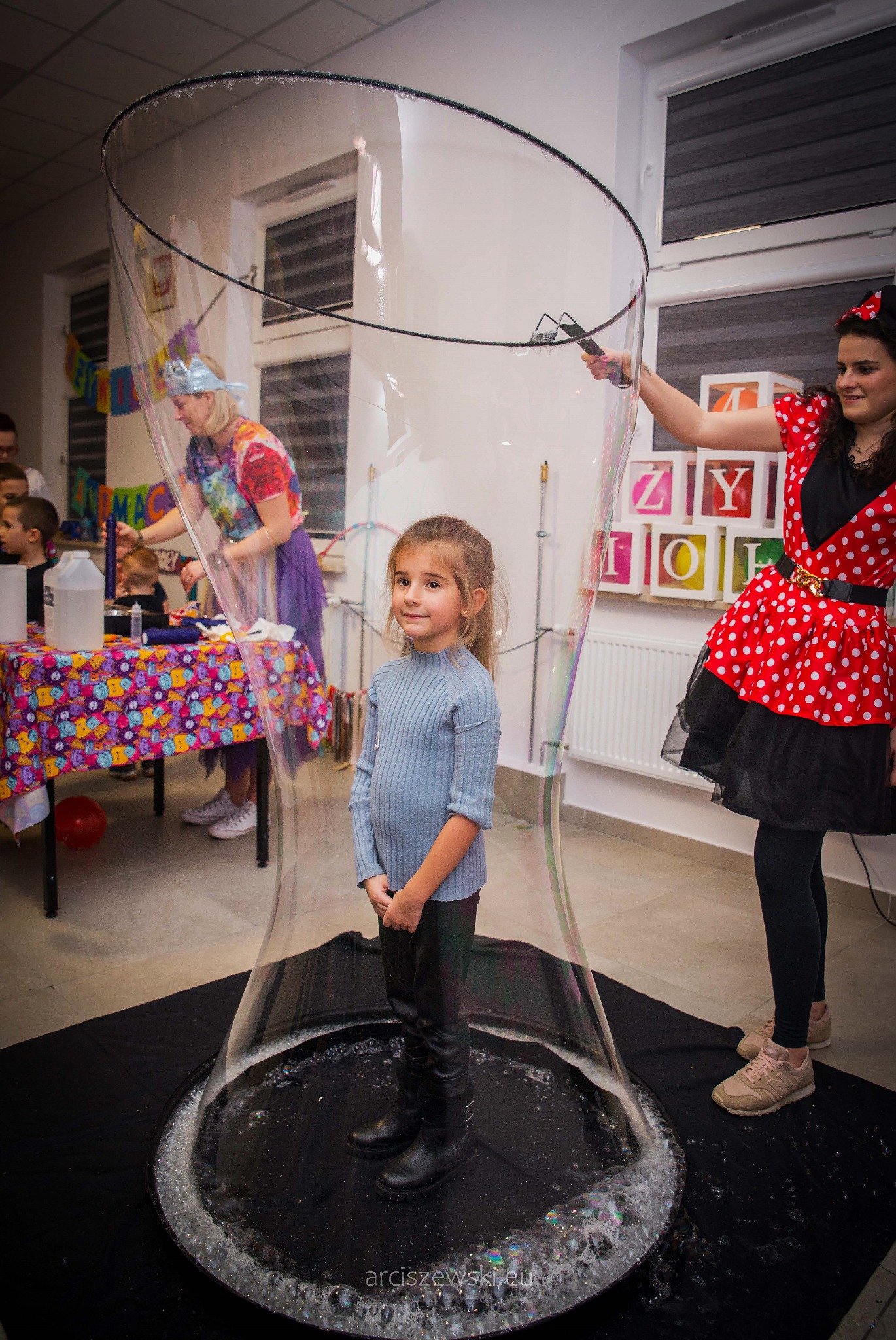 Dziewczynka stoi wewnątrz gigantycznej bańki mydlanej, tworzonej przez animatorkę w stroju Myszki Minnie. W tle stół z akcesoriami do baniek i bawiące się dzieci.