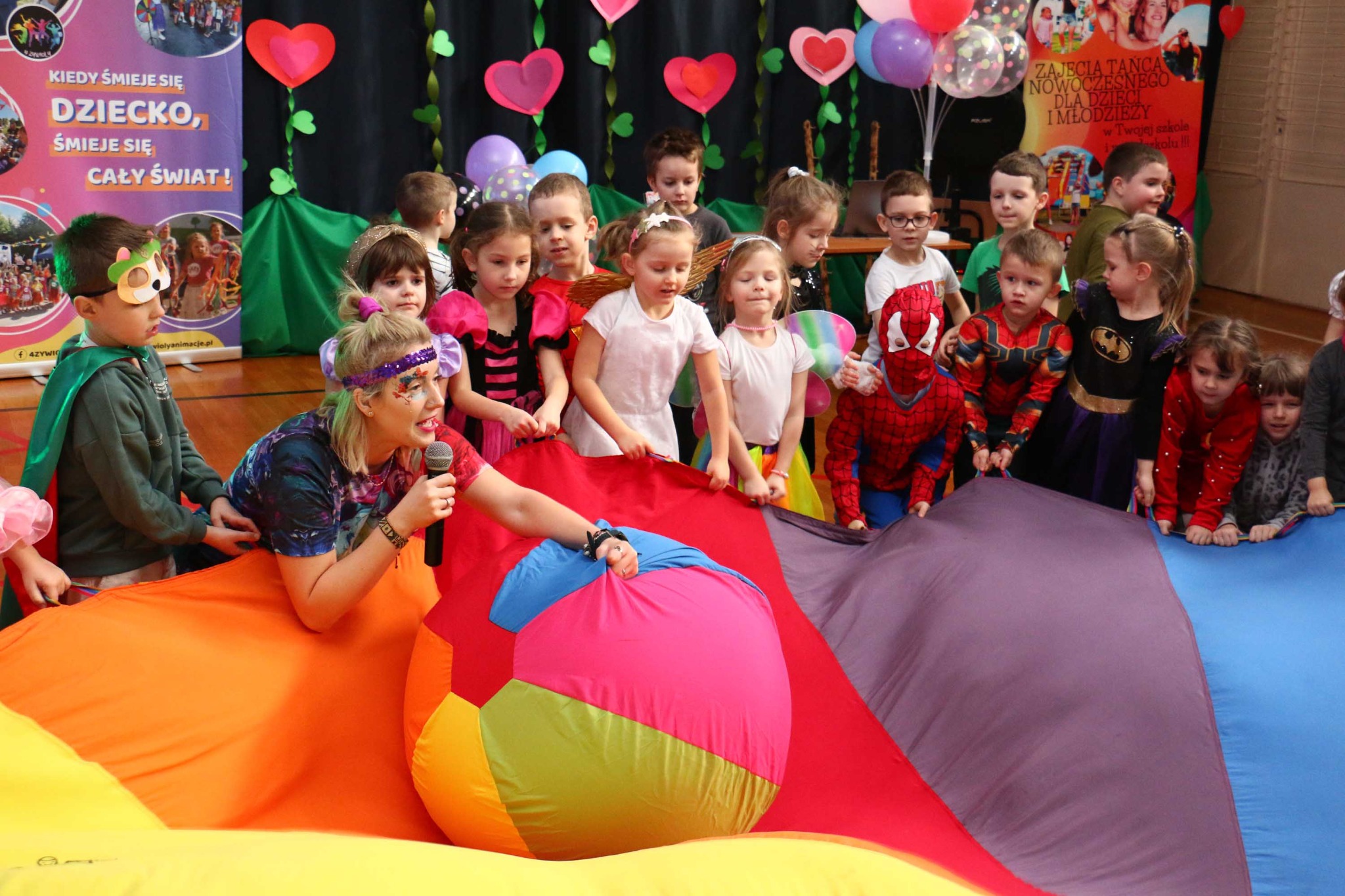 Animator z mikrofonem prowadzi zabawy z dziećmi trzymającymi kolorową chustę animacyjną. W tle dekoracje imprezowe i dzieci w kostiumach.