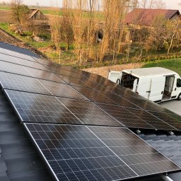 Pure Investments Group JĘDRZEJ KARCZEWSKI - Panele fotowoltaiczne na dachu domu jednorodzinnego, z białym vanem serwisowym w tle i wiejskim krajobrazem, uchwycone w ciepłym świetle słonecznym.