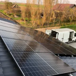 Panele fotowoltaiczne na dachu domu jednorodzinnego, z białym vanem serwisowym w tle i wiejskim krajobrazem, uchwycone w ciepłym świetle słonecznym.