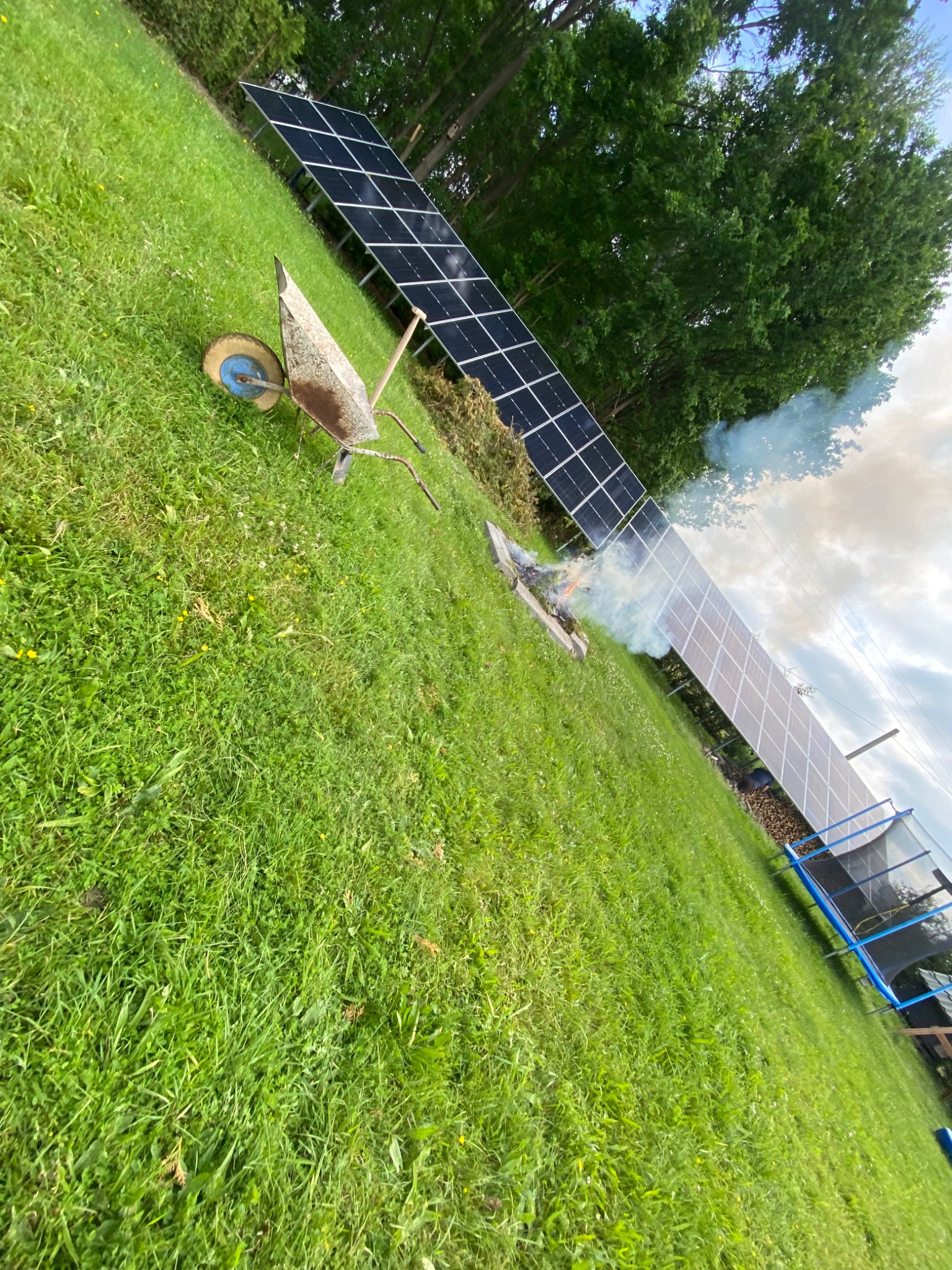 Panele słoneczne na trawie, obok płonie ognisko, a w pobliżu stoi taczka. Nietypowe ujęcie instalacji fotowoltaicznej w ogrodzie.